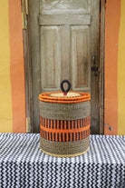 Handwoven Bolga laundry basket with coiled rim and lid in natural straw with a bold orange banded stripe.
