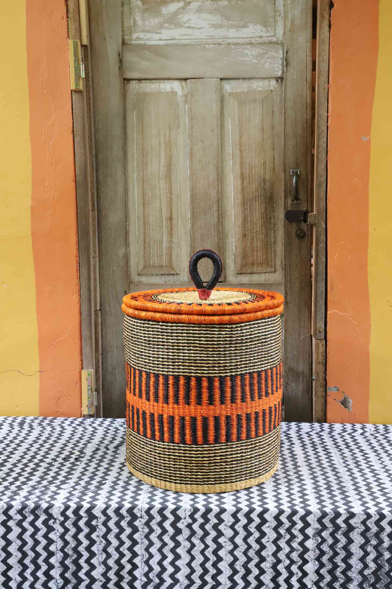 Handwoven Bolga laundry basket with coiled rim and lid in natural straw with a bold orange banded stripe.