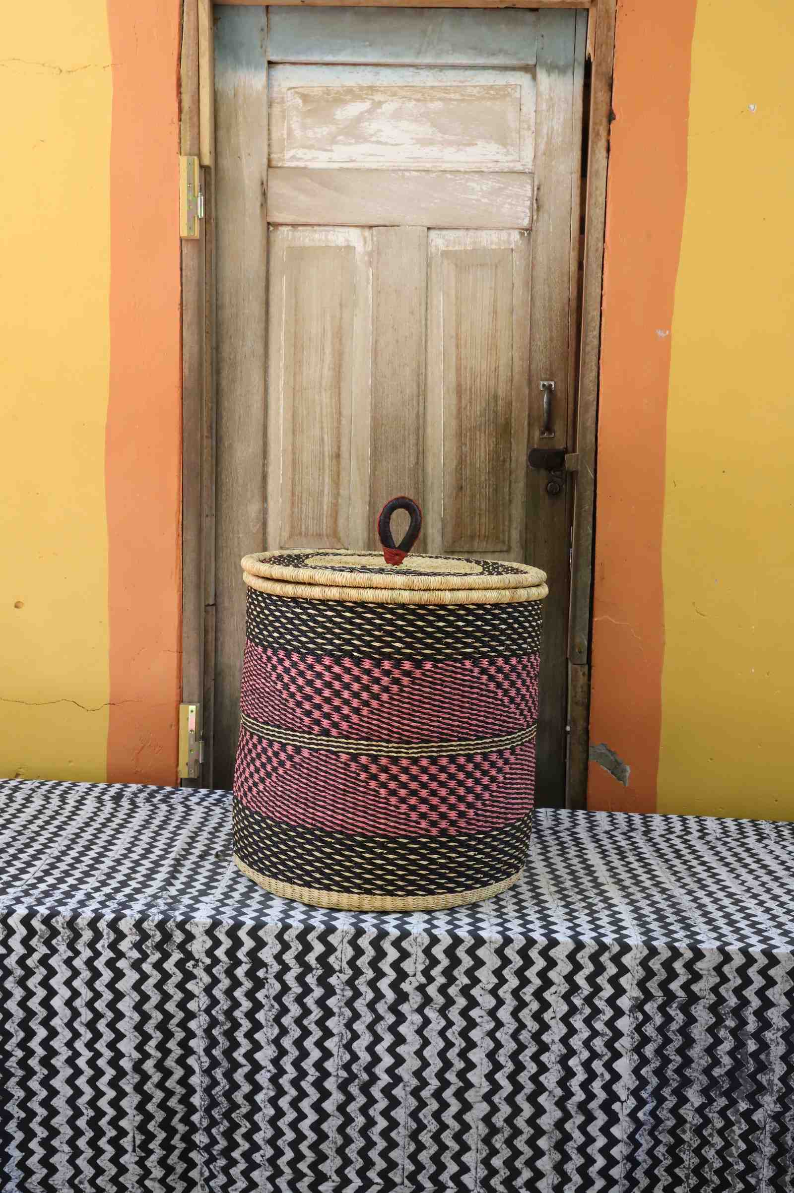 Handwoven Bolga laundry basket with coiled rim and lid in natural straw with red-pink checkered central band.