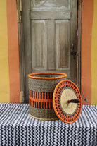 Handwoven Bolga laundry basket with coiled rim and lid removed, natural straw body with orange band.