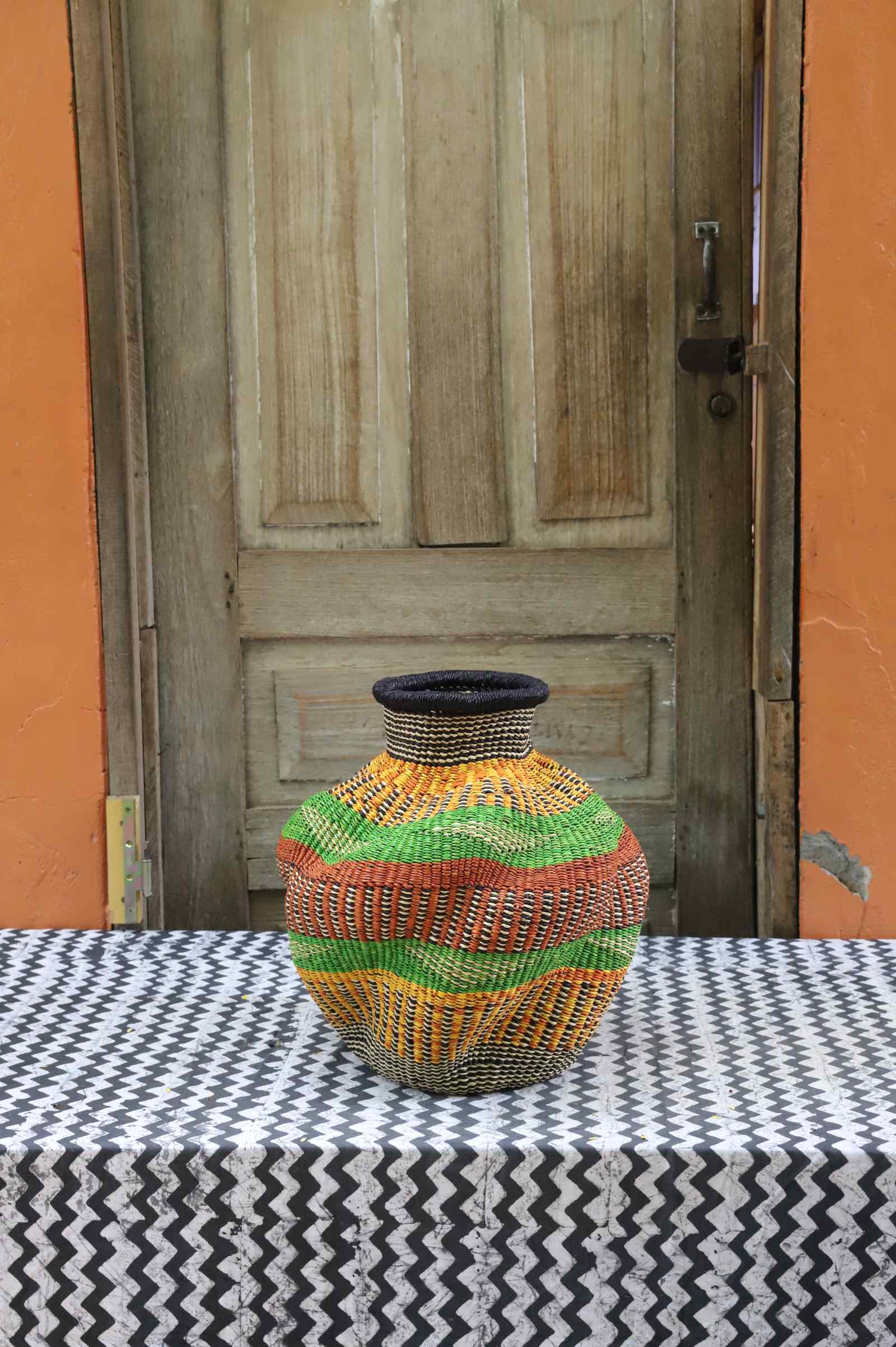 Handwoven Bolga art basket in an urn shape with dark rim, green, orange and yellow banded stripes.