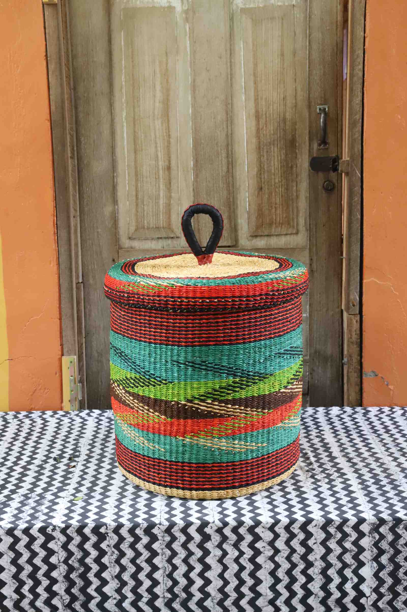 Lidded laundry basket in rust and teal wave-patterned weave, styled on a chevron cloth by a wooden door.