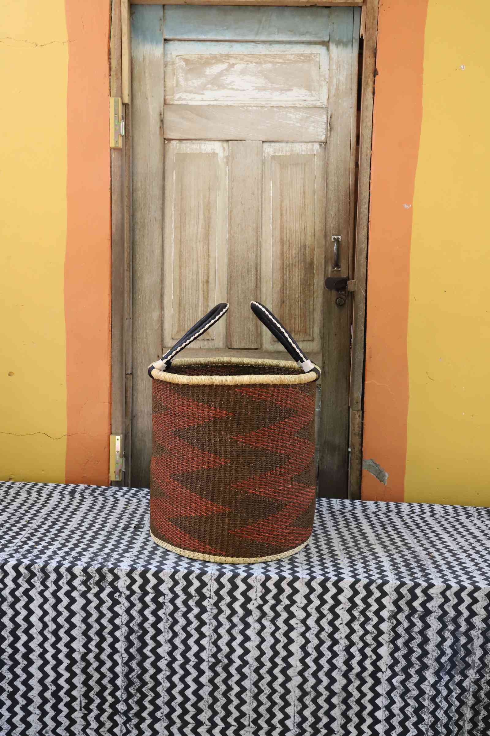 Medium handwoven open-top Bolga laundry basket in rust-orange with dark diagonal zigzag stripes.