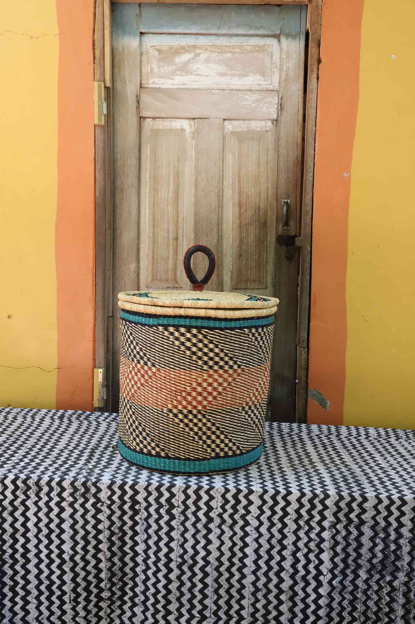 Handwoven Bolga laundry basket with coiled rim and lid in natural straw with orange and dark banded stripes.