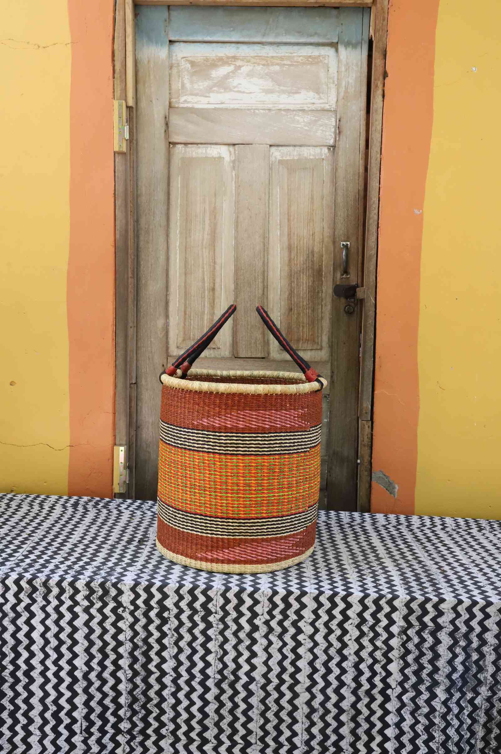Medium handwoven open-top Bolga laundry basket in natural straw with orange, yellow and red fine bands.