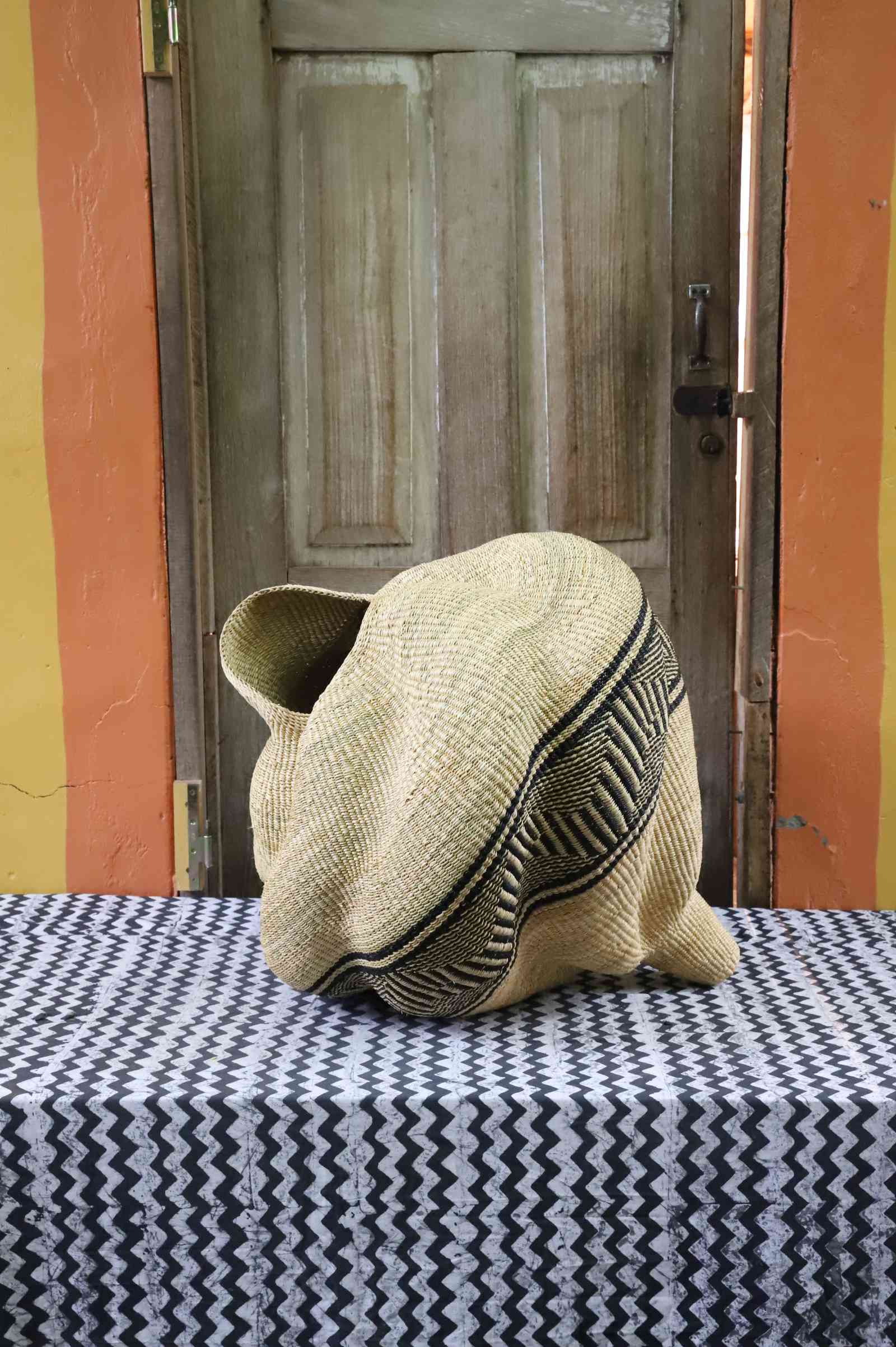 Sculptural spiral-form Bolga basket in natural straw with a black geometric band, styled on a patterned tablecloth.