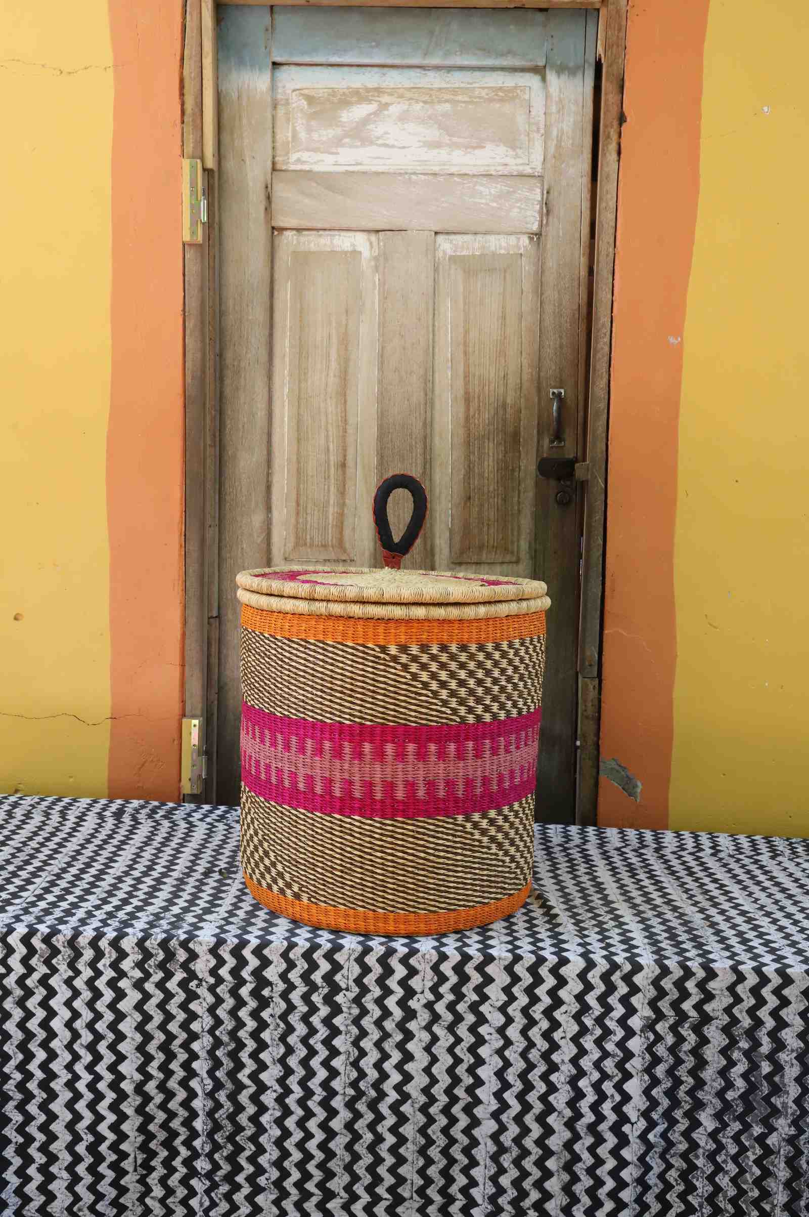 Handwoven Bolga laundry basket with coiled rim and lid in natural straw with pink and orange central bands.