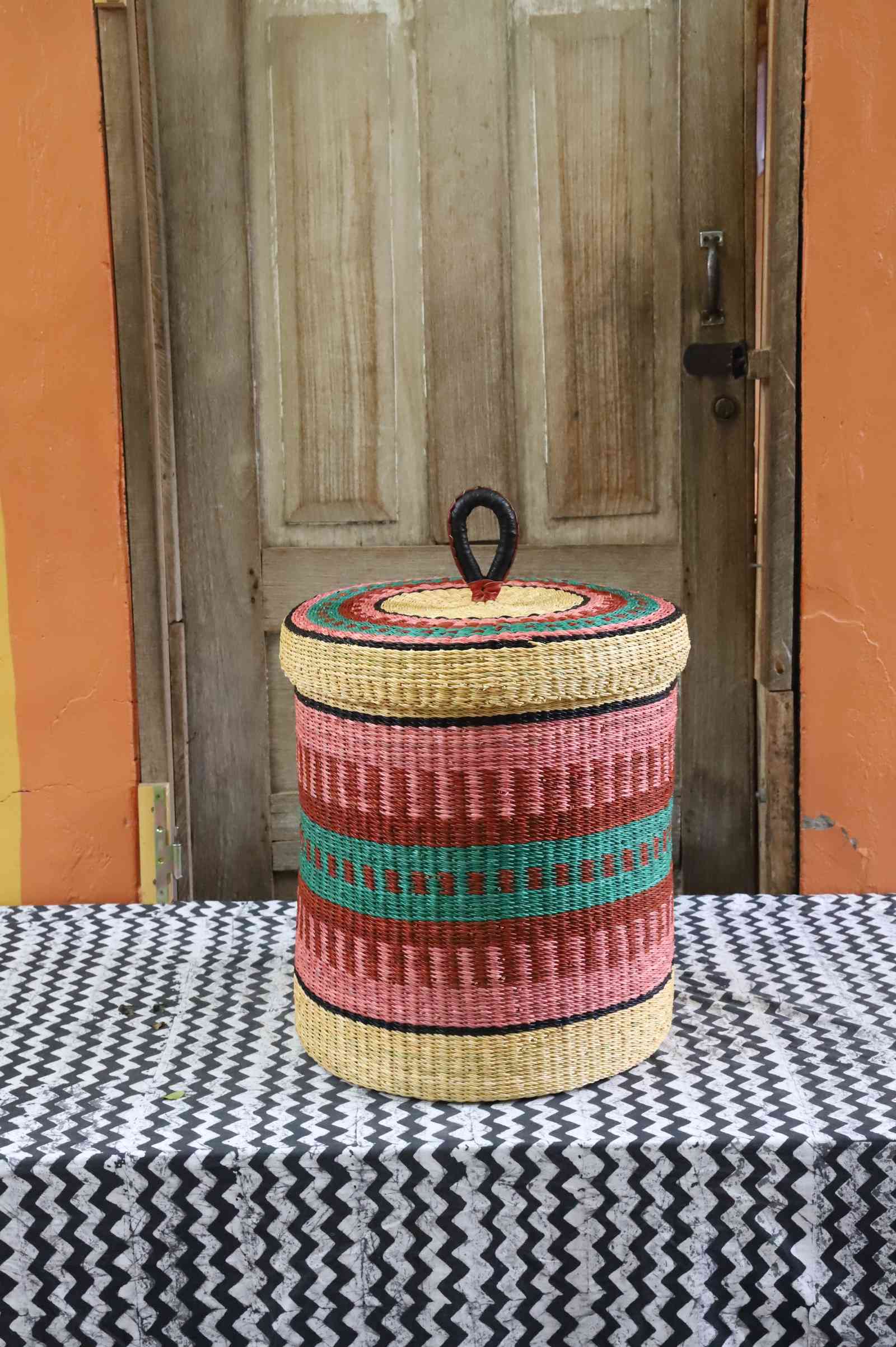 Lidded laundry basket in natural straw with pink and teal striped banding, styled on a chevron cloth by a wooden door.