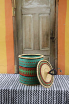 Handwoven Bolga laundry basket with lid removed, showing teal, red and green banded body and lid.