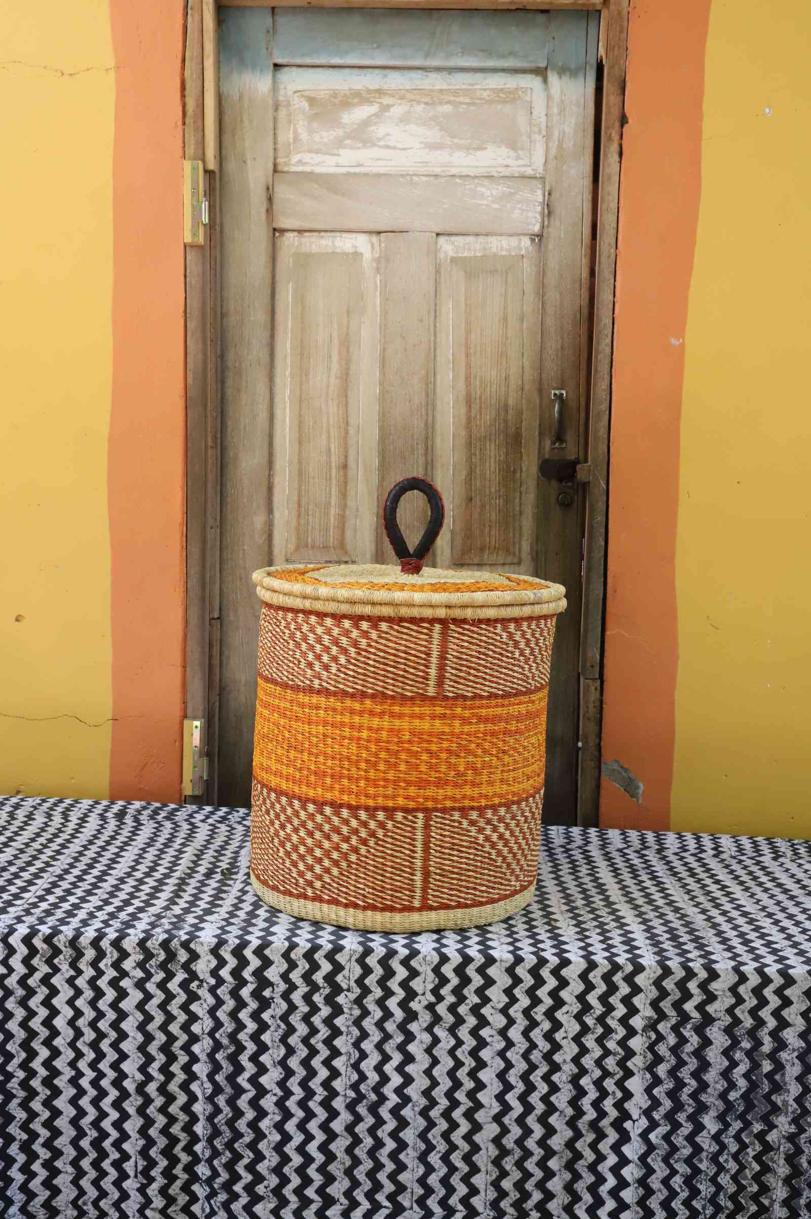 Handwoven Bolga laundry basket with coiled rim and lid in natural straw with an orange central band.