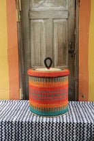 Handwoven Bolga laundry basket with lid in orange elephant grass with teal horizontal stripes.