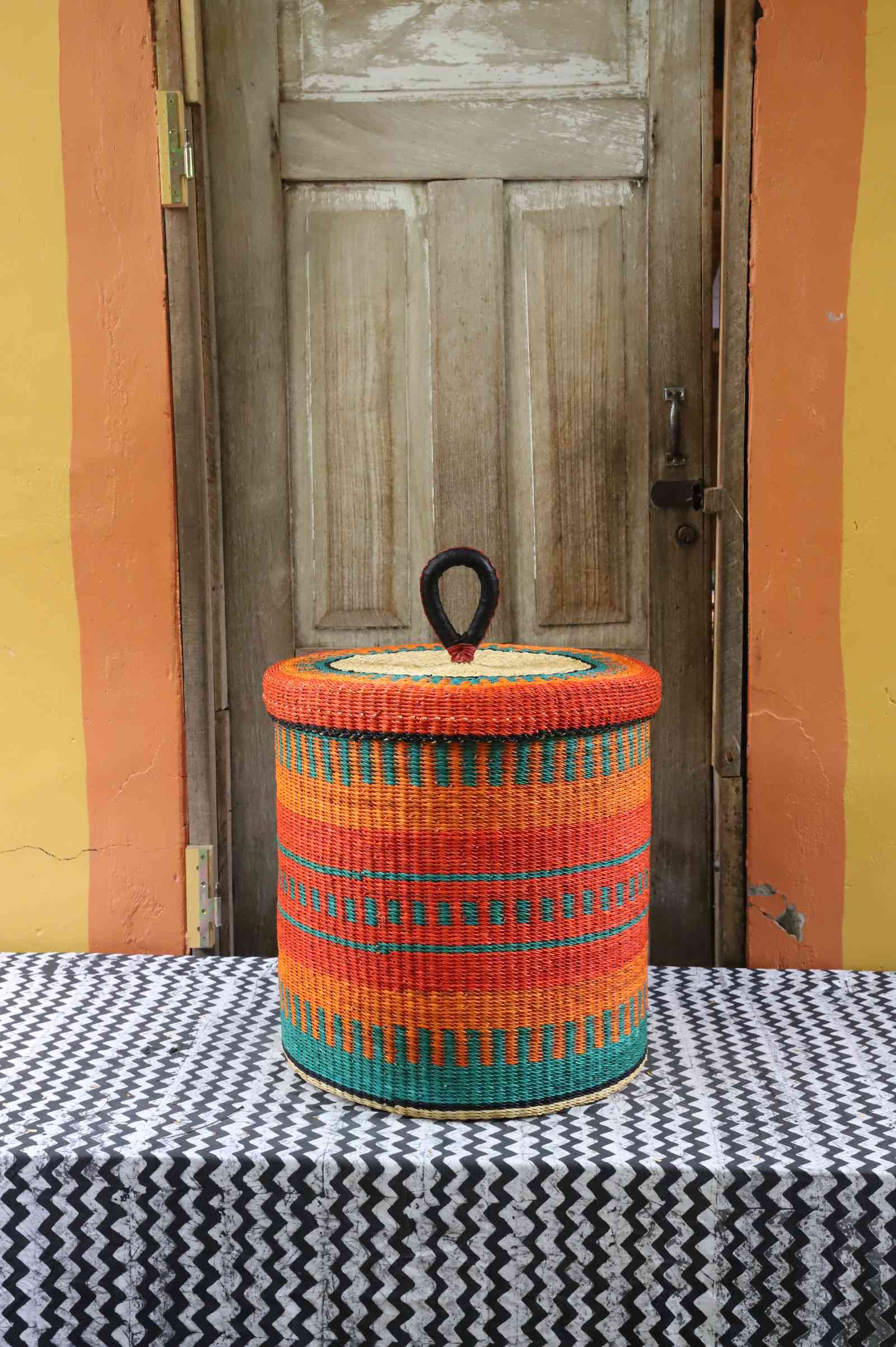 Handwoven Bolga laundry basket with lid in orange elephant grass with teal horizontal stripes.