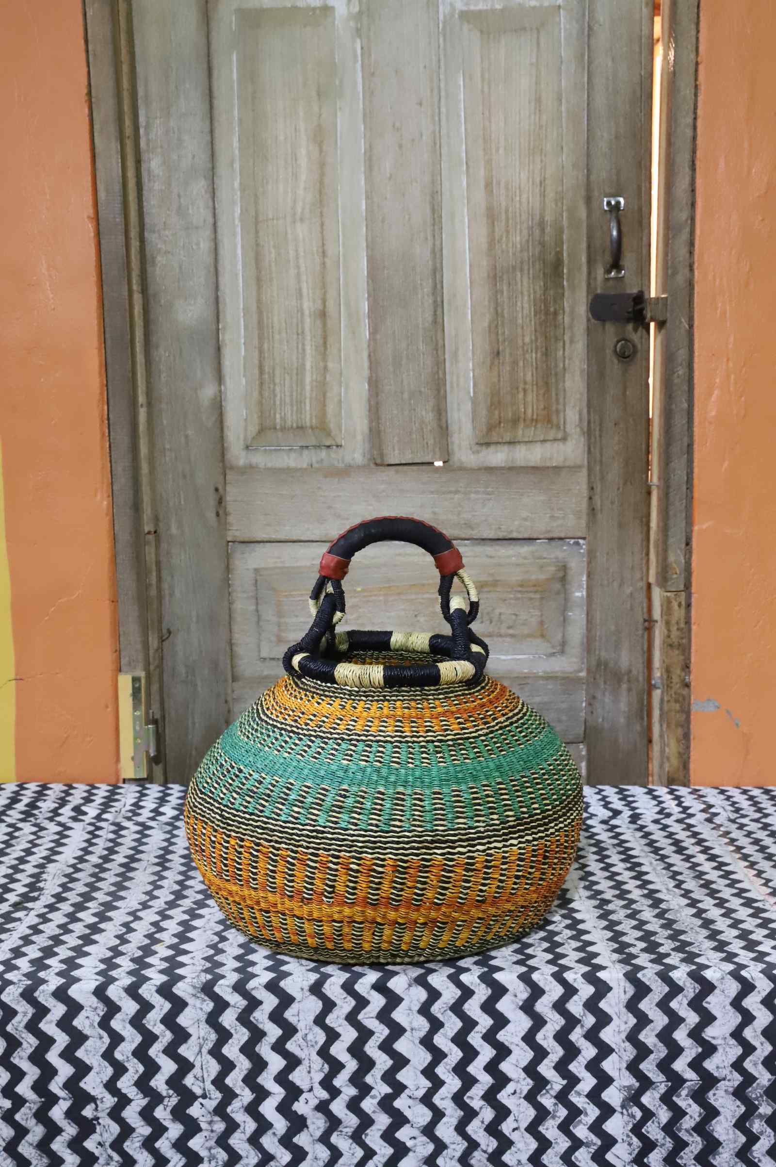 Round Bolga pot basket in orange and green stripes with a leather-wrapped arched handle, on a tablecloth.