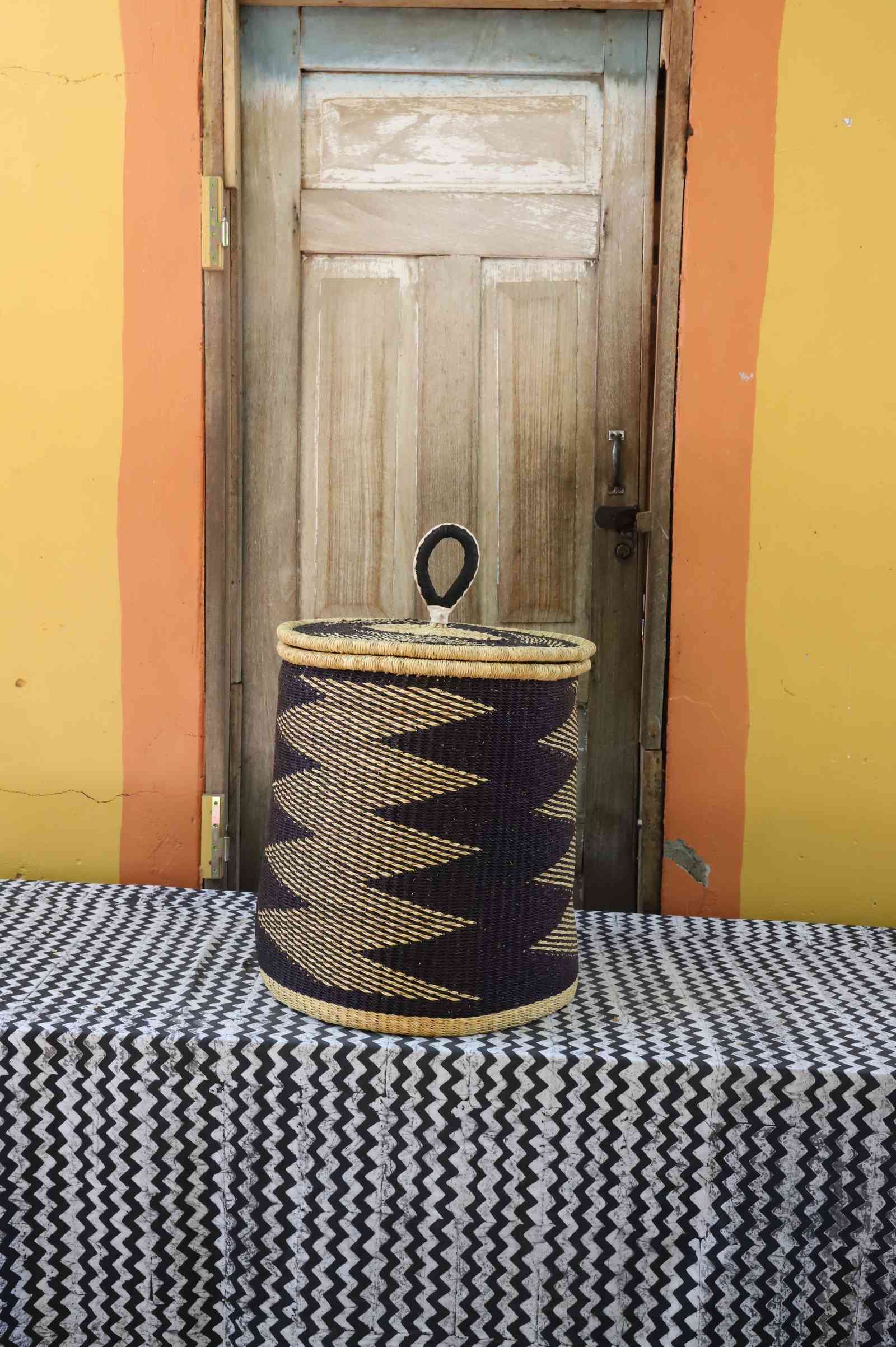 Handwoven Bolga laundry basket with coiled rim and lid in natural straw with a bold black zigzag lower band.