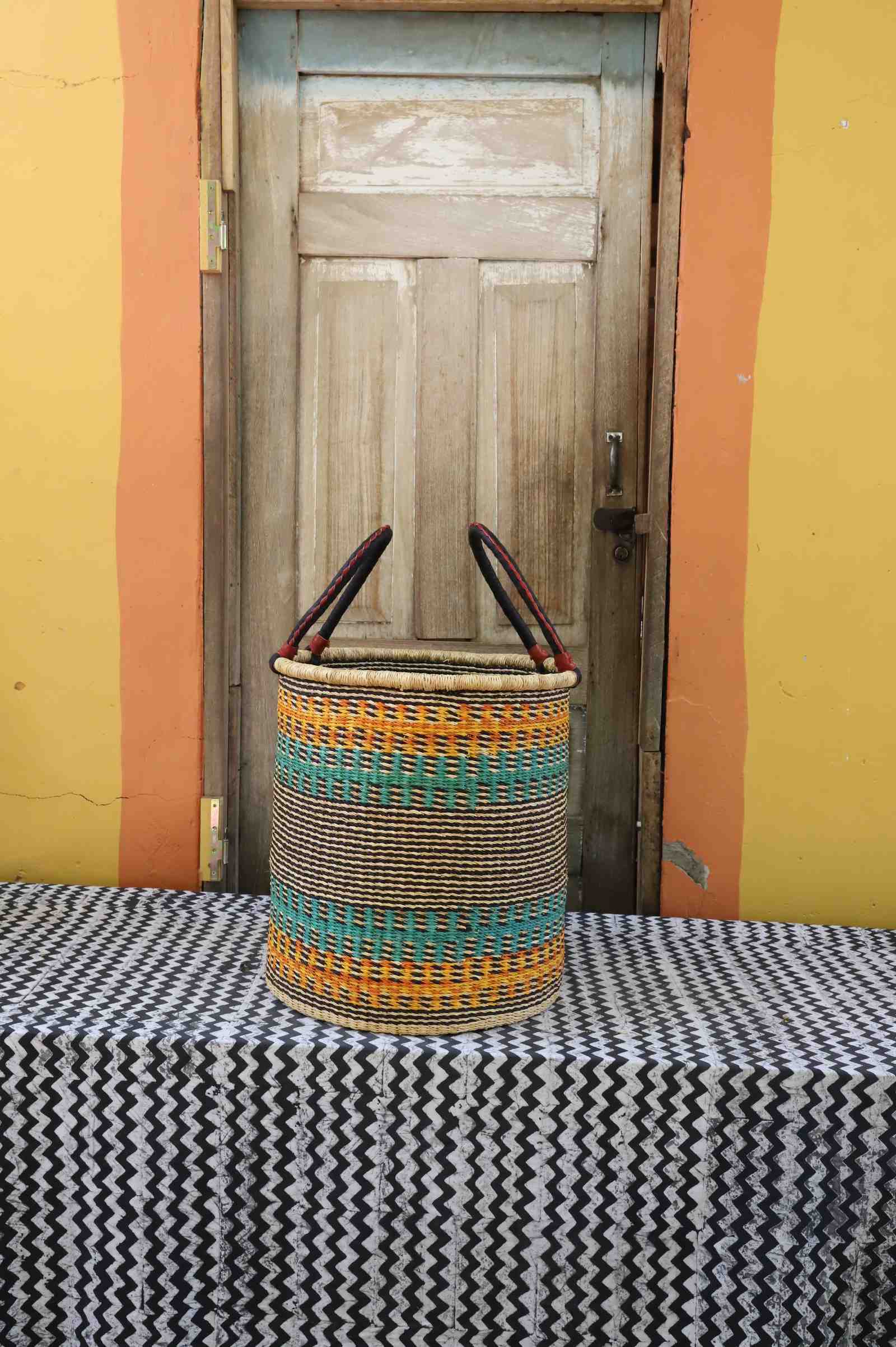 Medium handwoven open-top Bolga laundry basket in natural straw with orange, teal and green banded stripes.