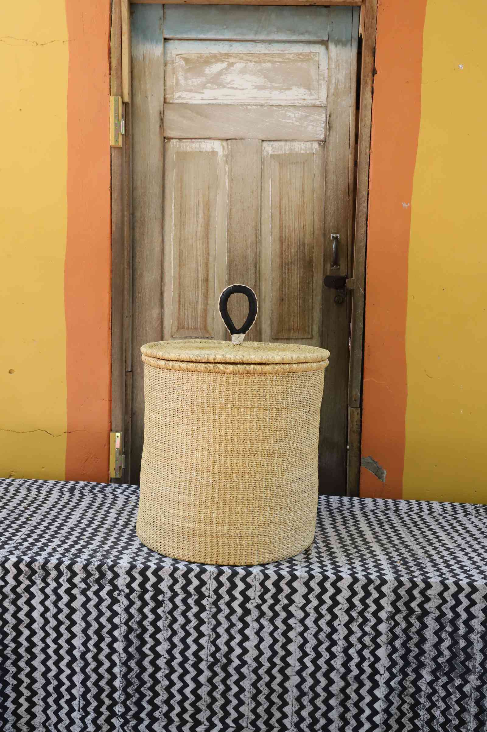 Handwoven Bolga laundry basket with coiled rim and lid in plain natural straw with a dark rope handle.