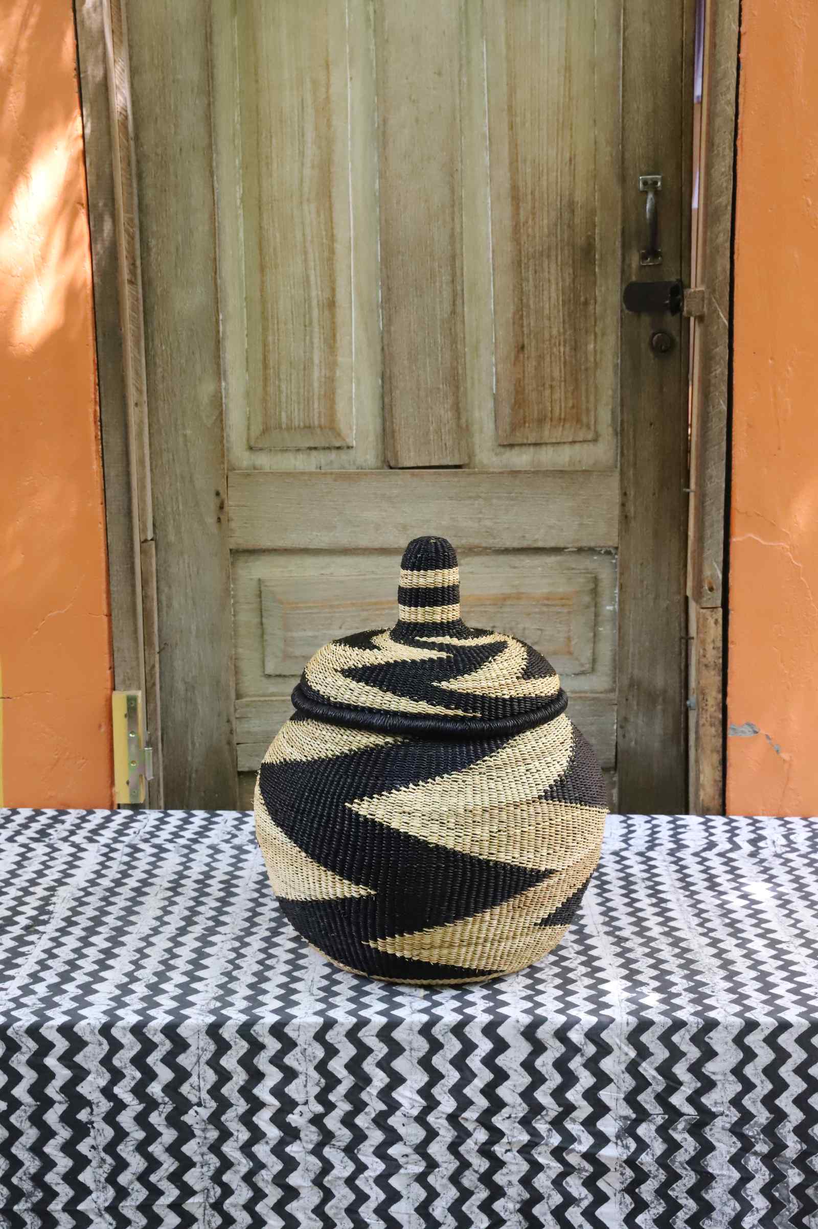 Lidded Bolga storage basket with a bold black and natural zigzag pattern and a domed fitted lid, on a tablecloth.