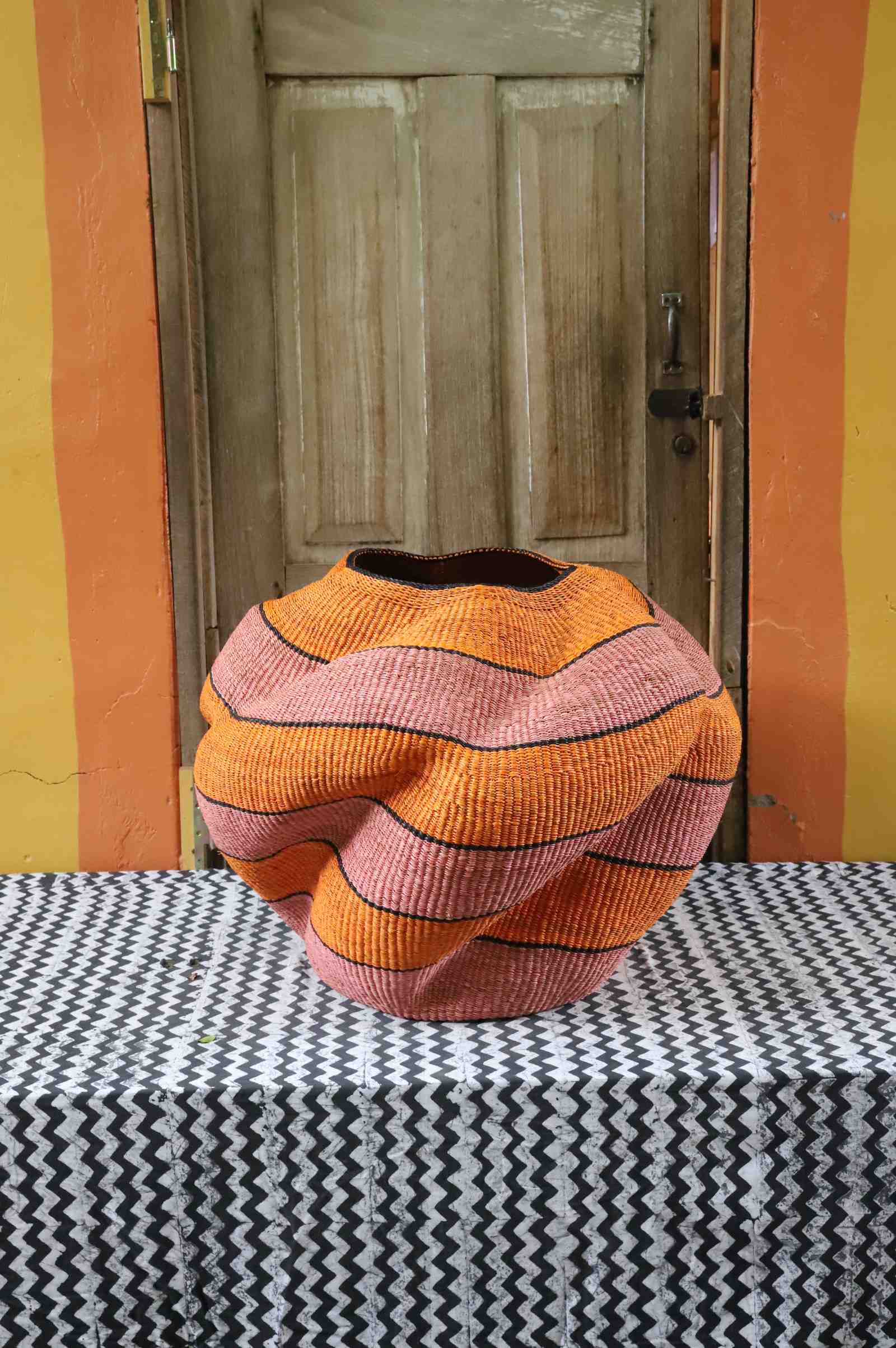 Large nest-shaped Bolga art basket in pink and rust wavy braided weave, styled on a chevron cloth by a wooden door.