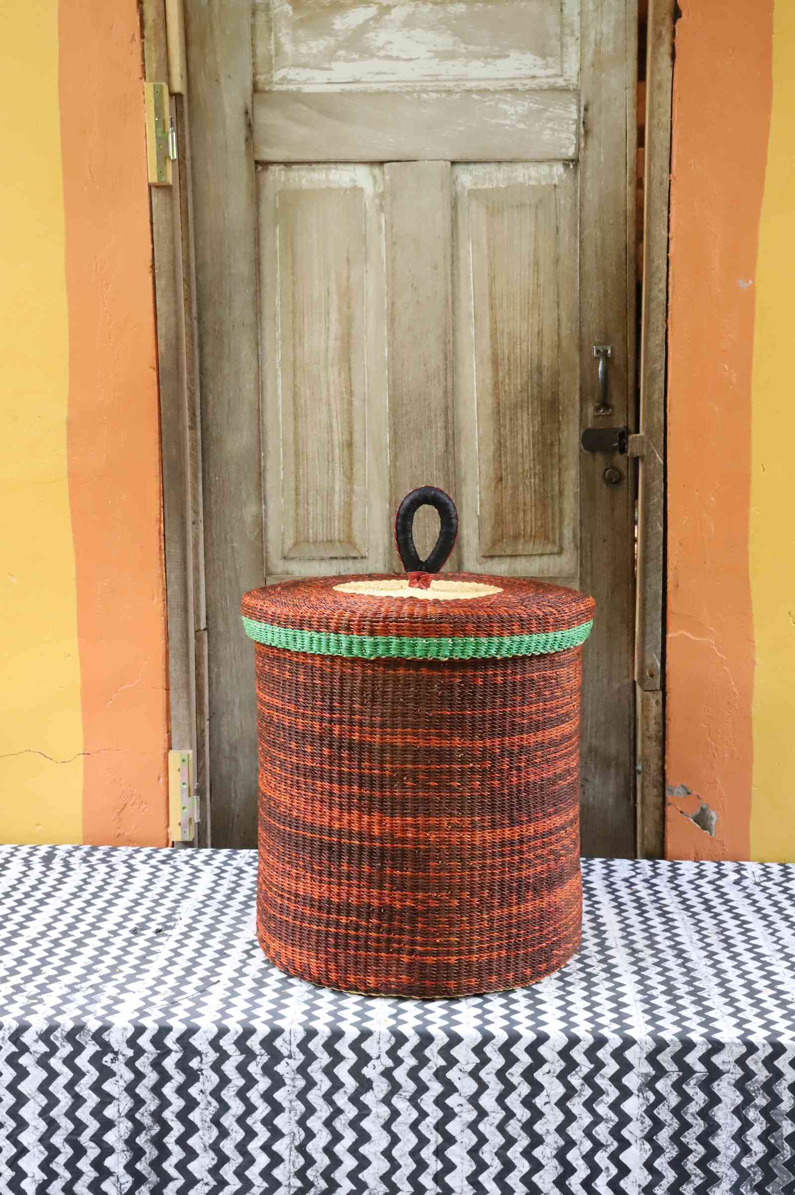 Handwoven Bolga laundry basket with lid in rust-orange elephant grass with a green banded rim.