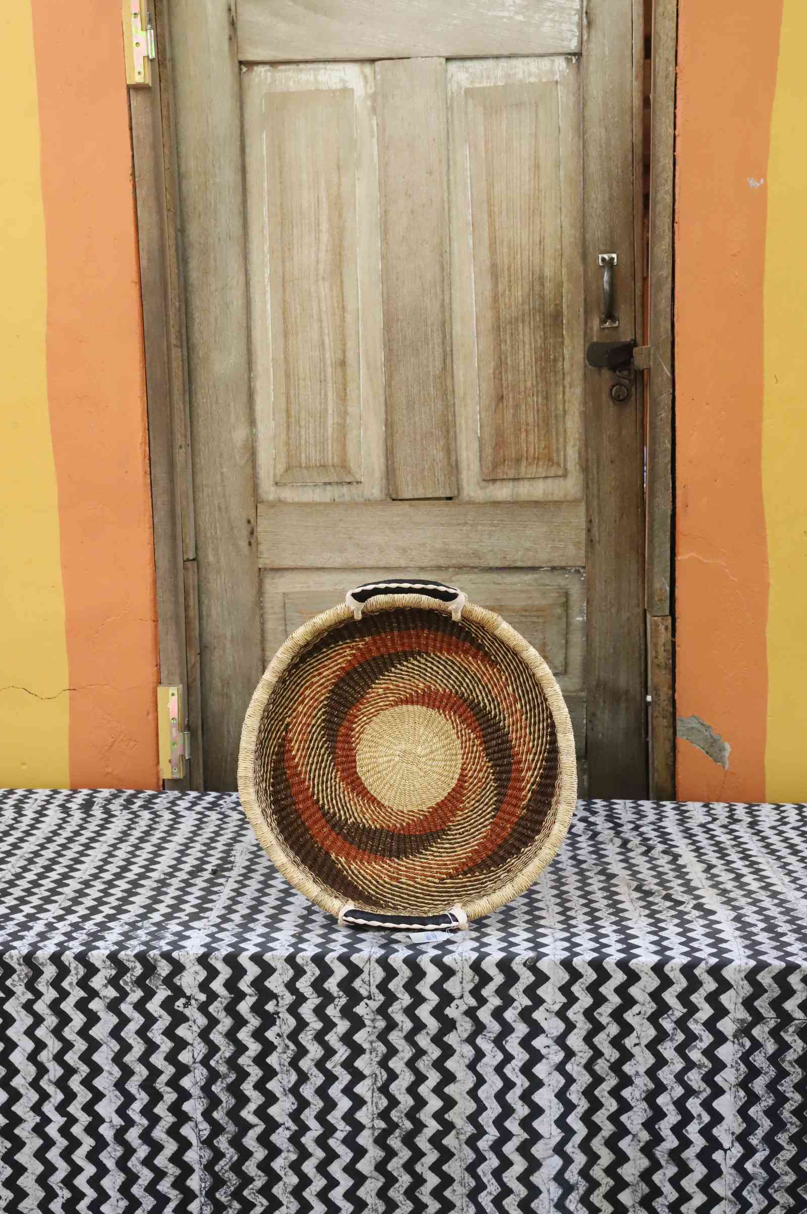 Handwoven Bolga wall tray disc in natural straw with rust-red pinwheel swirl and yellow centre.