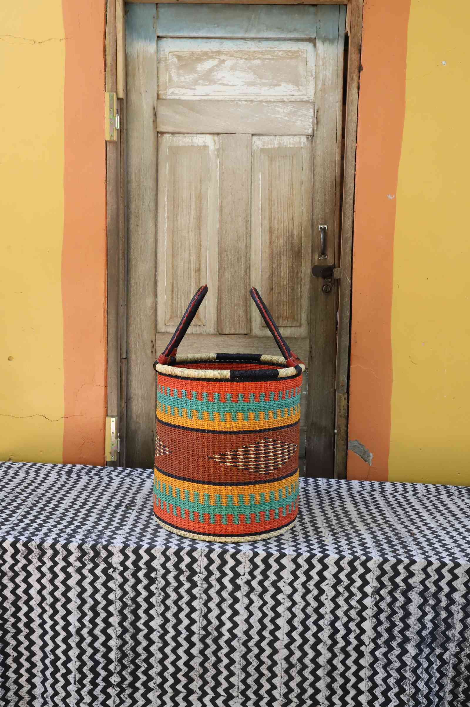 Small handwoven open-top Bolga laundry basket in dyed orange with green and red banded stripes.