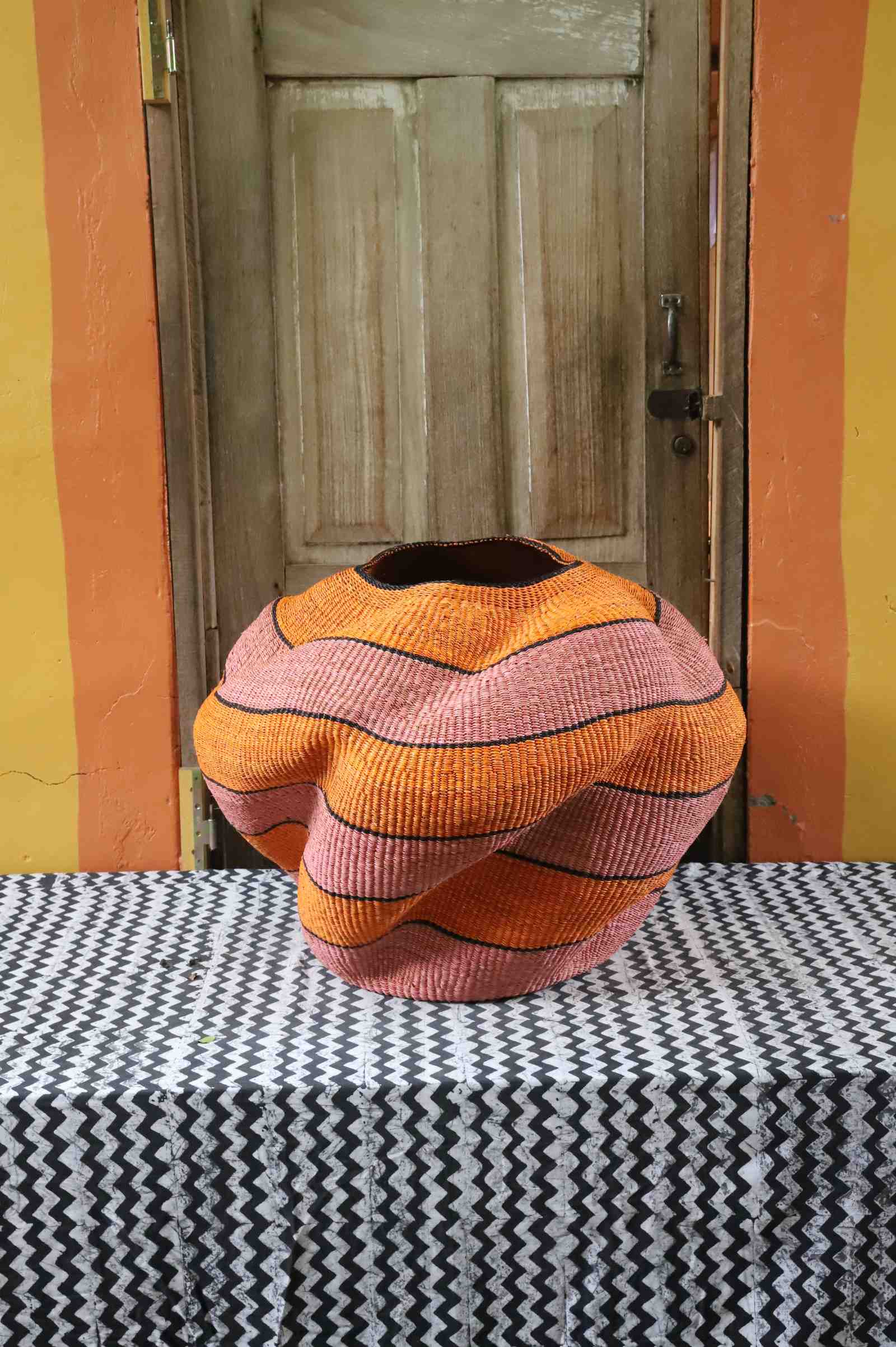 Large nest-shaped Bolga art basket in pink and rust wavy braided weave, front view on a chevron cloth.