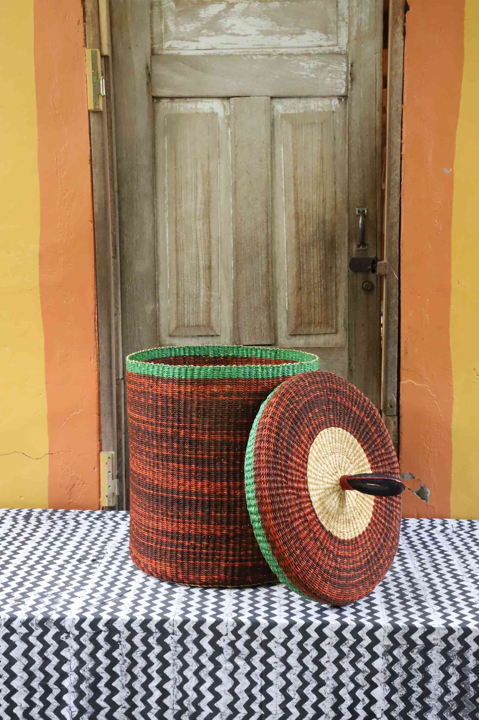 Handwoven Bolga laundry basket with lid removed, showing rust-orange body with green rim and matching lid.