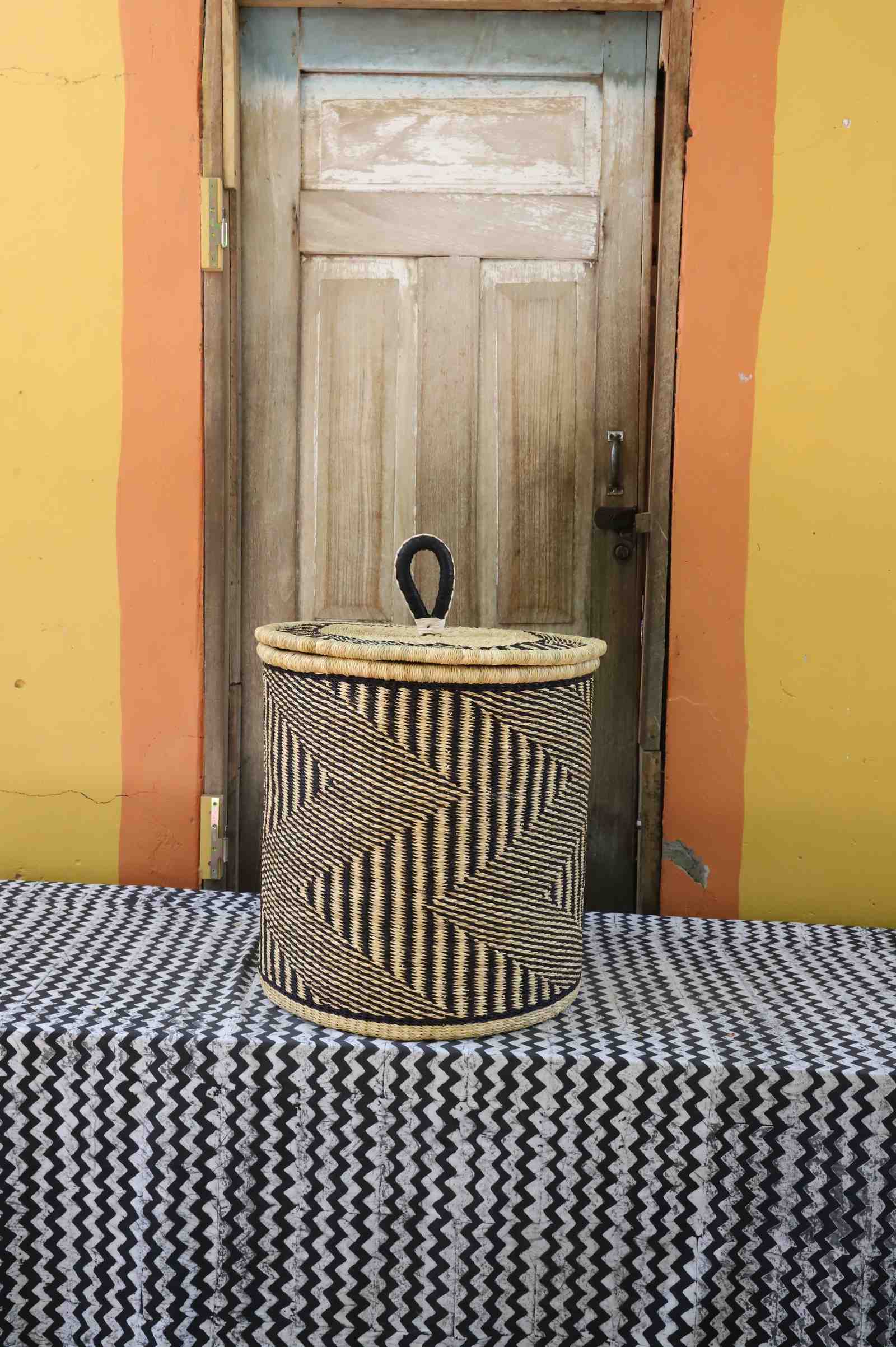Handwoven Bolga laundry basket with coiled rim and lid in natural straw with dark diagonal wave stripes.