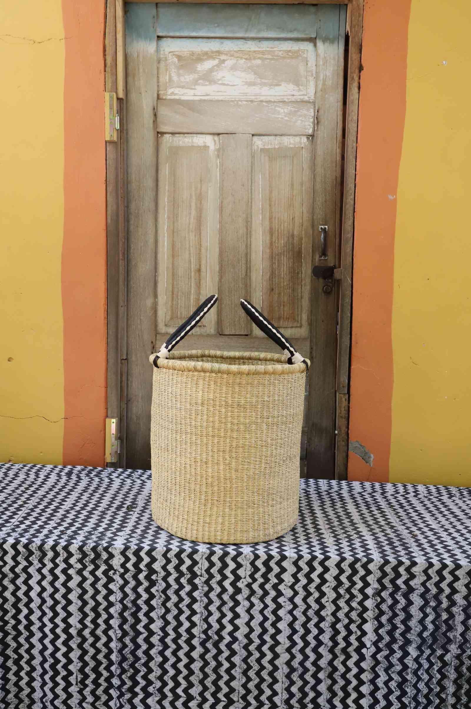 Small handwoven open-top Bolga laundry basket in plain natural straw with twin looped handles.