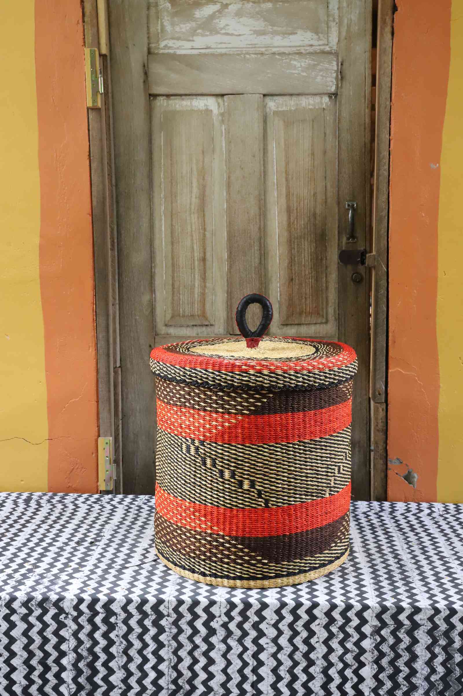 Handwoven Bolga laundry basket with lid in natural straw with red-orange stripes and a black diamond weave band.