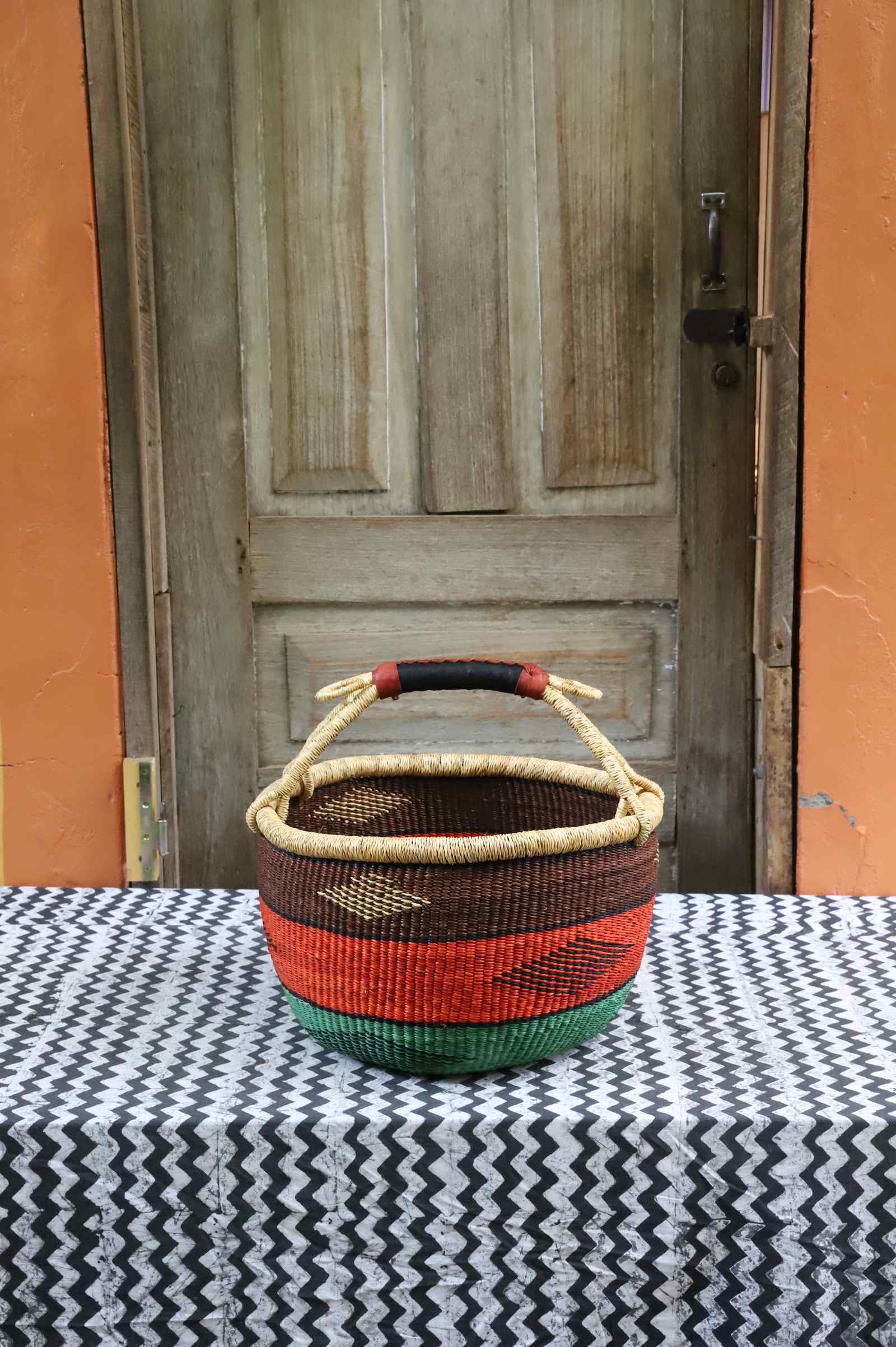 Round Bolga shopping basket with a leather-wrapped handle, striped in brown, orange and green, on a tablecloth.