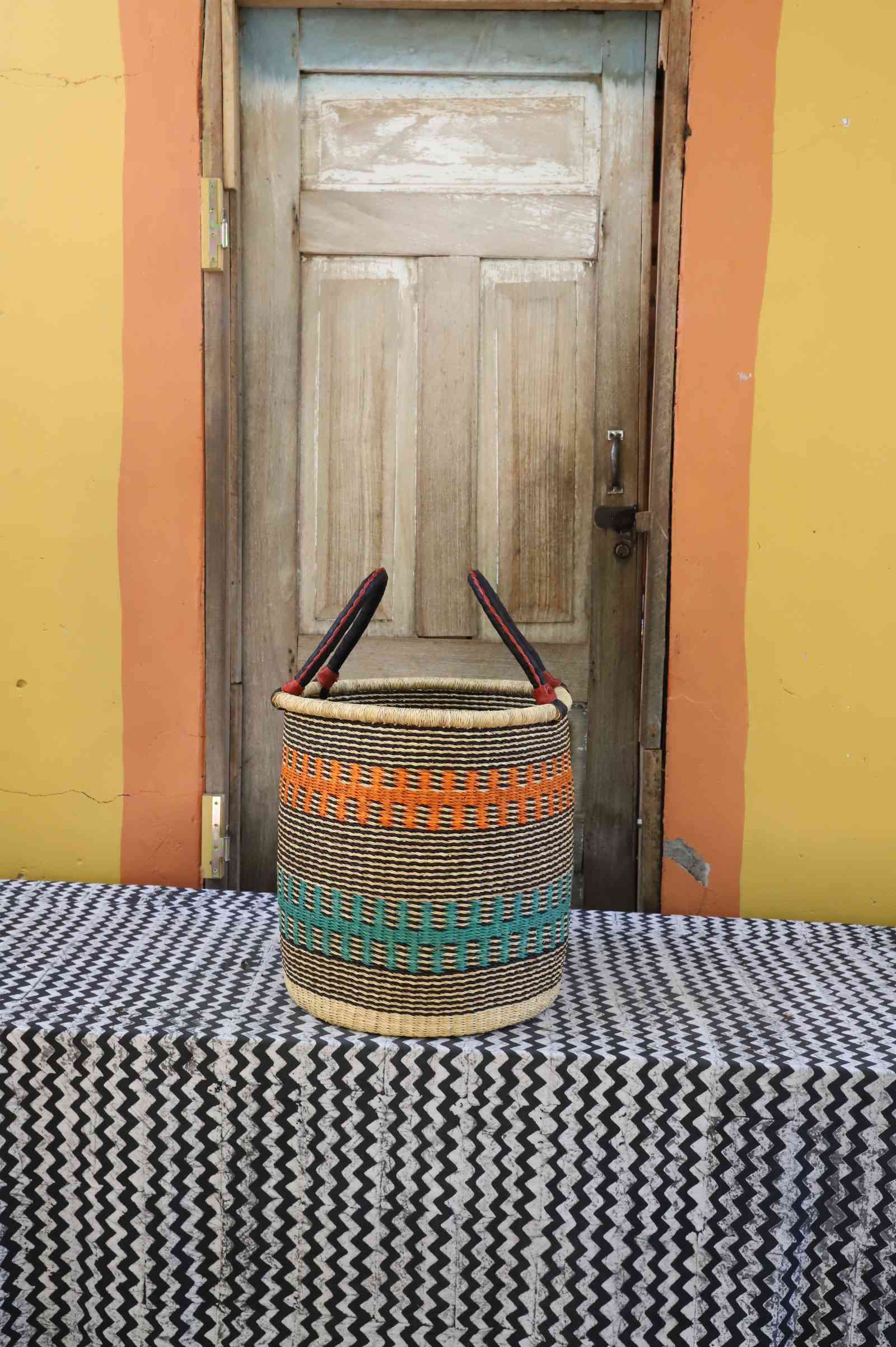 Small handwoven open-top Bolga laundry basket in natural straw with orange and teal banded stripes.