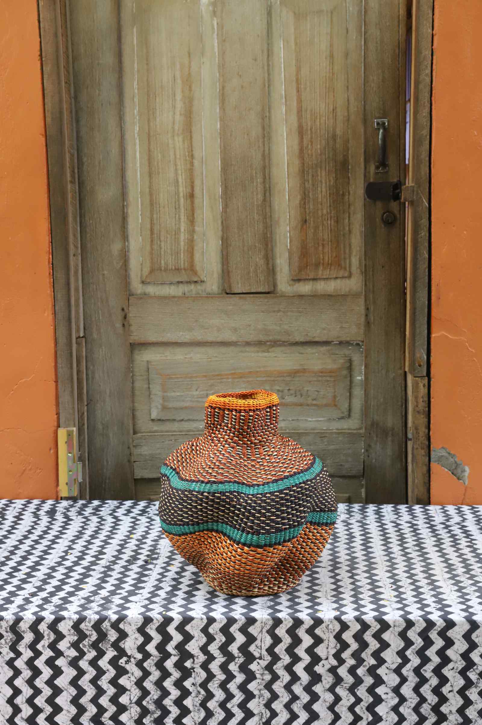 Handwoven Bolga art basket in a rounded pot shape, natural straw with orange rim and a teal and orange central wave band.