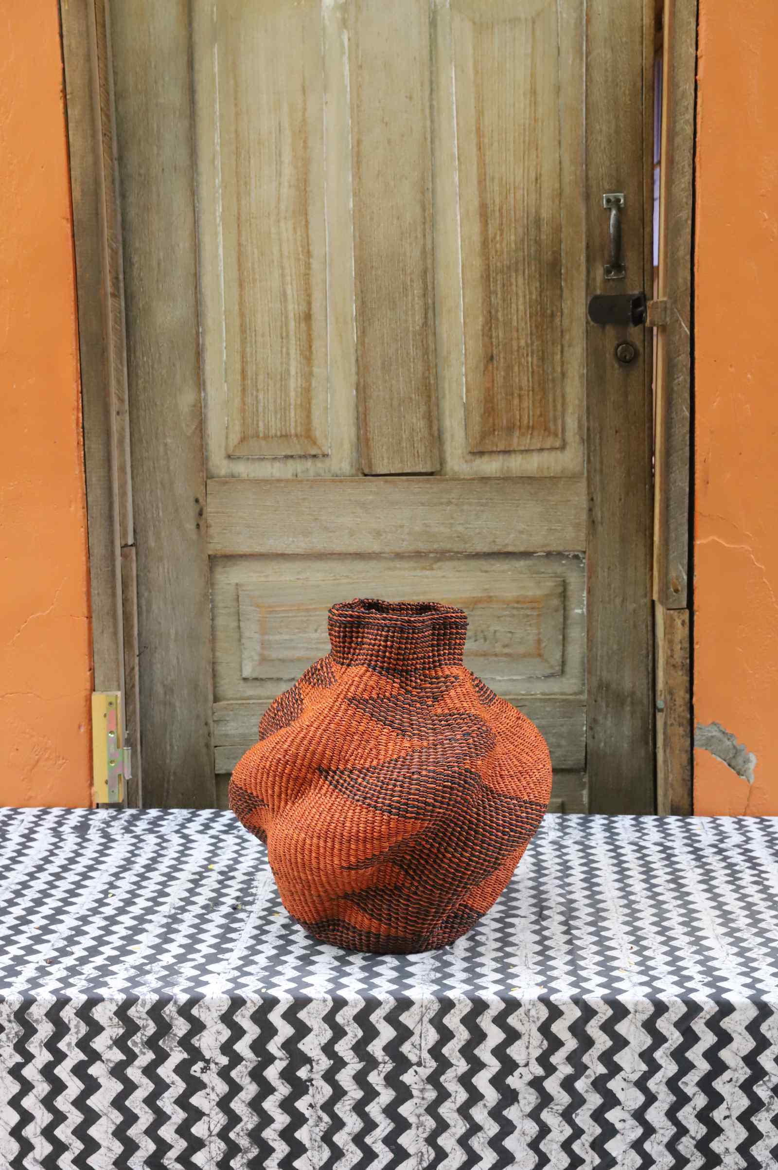 Handwoven Bolga art basket in a rounded pot shape, dyed rust-orange with a black triangular arrow motif.