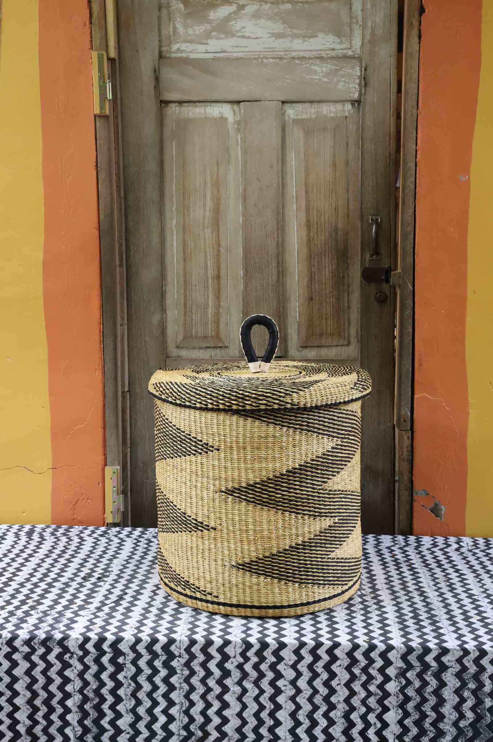 Handwoven Bolga laundry basket with lid in natural straw with a black zigzag pattern and looped lid handle.