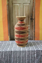 Tall vase-shaped Bolga art basket in natural straw with a brown and rust diamond pattern, styled on a chevron cloth by a wooden door.