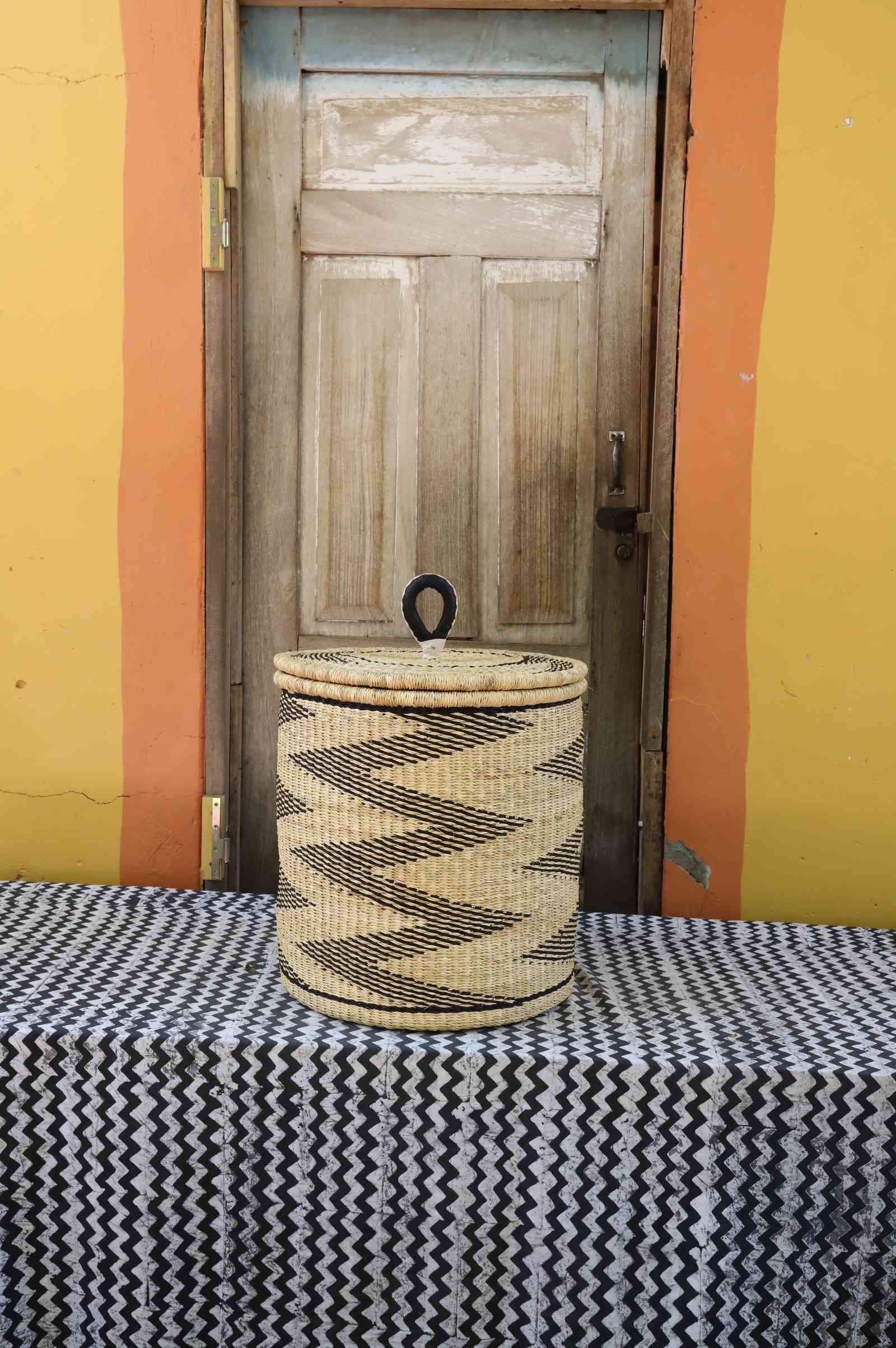Handwoven Bolga laundry basket with coiled rim and lid in natural straw with a bold black zigzag wave motif.
