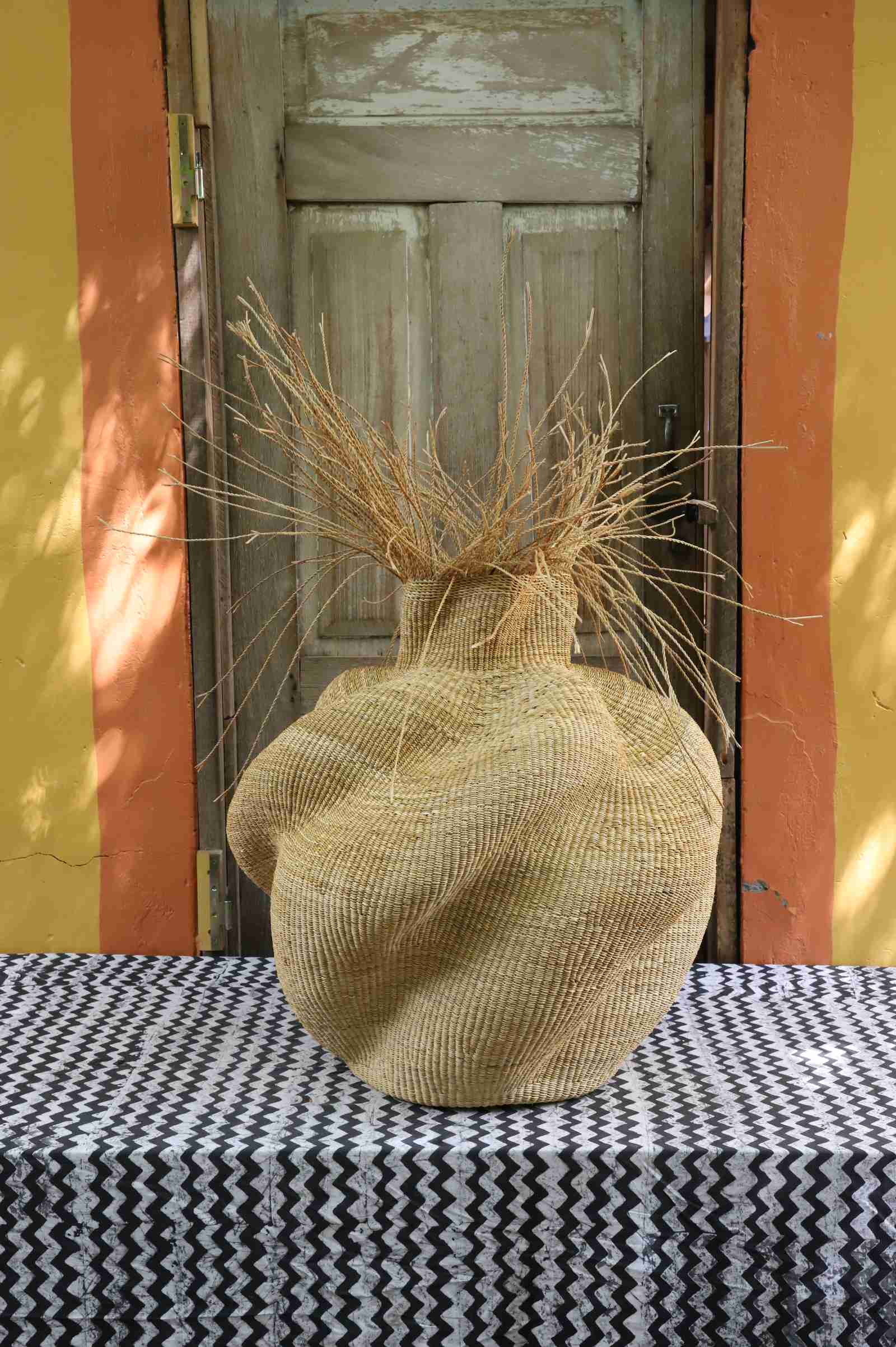 Bulbous Bolga basket in natural straw, its narrow neck topped with loose straw fronds, styled on a patterned tablecloth.
