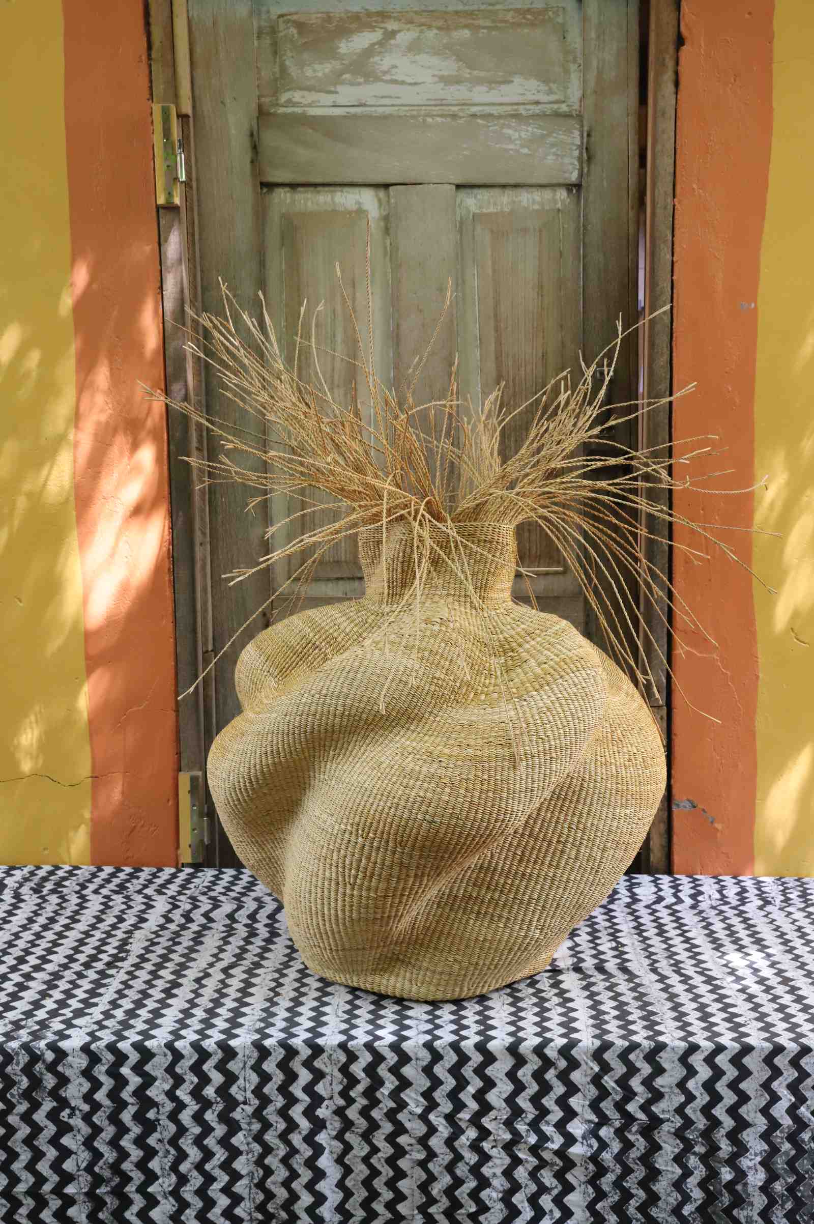 Round Bolga basket in natural straw with a bulbous woven body and loose straw fronds at the neck.