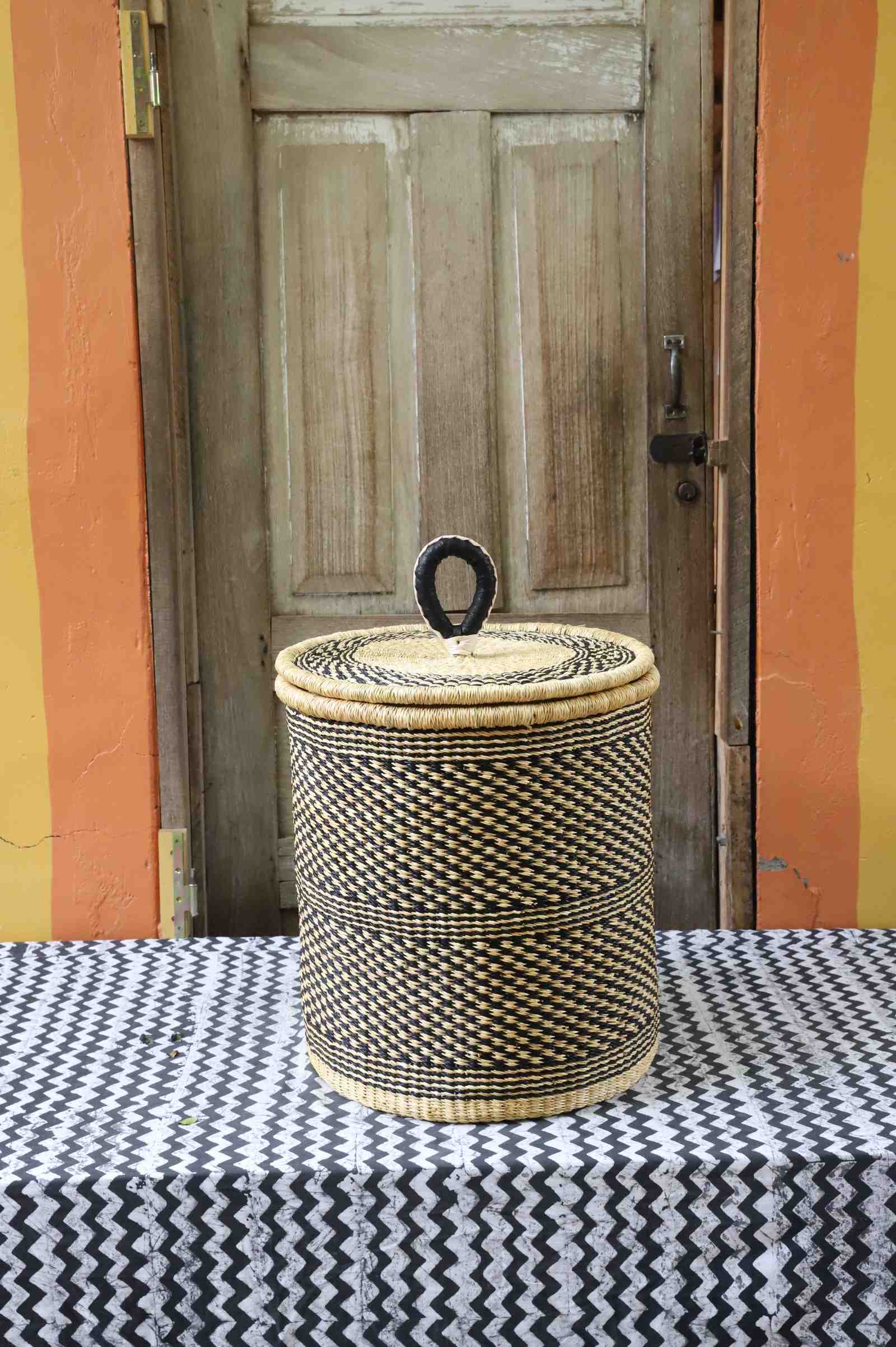 Coiled-rim lidded laundry basket in a black and tan speckled weave, styled on a chevron cloth by a wooden door.