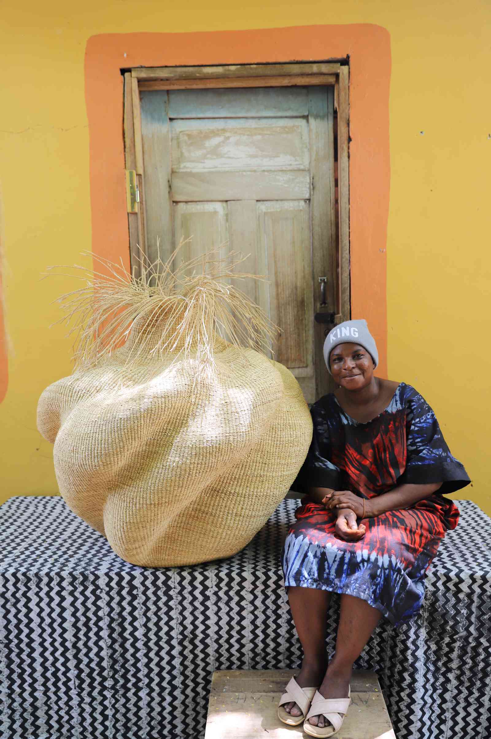 Handwoven art basket in natural straw with organic lobed body and dried grass stems, beside a woman seated in Bolgatanga.
