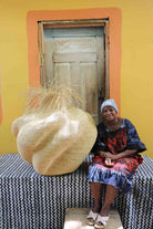 Handwoven art basket in natural straw with organic lobed body and dried grass stems, beside a woman seated in Bolgatanga.