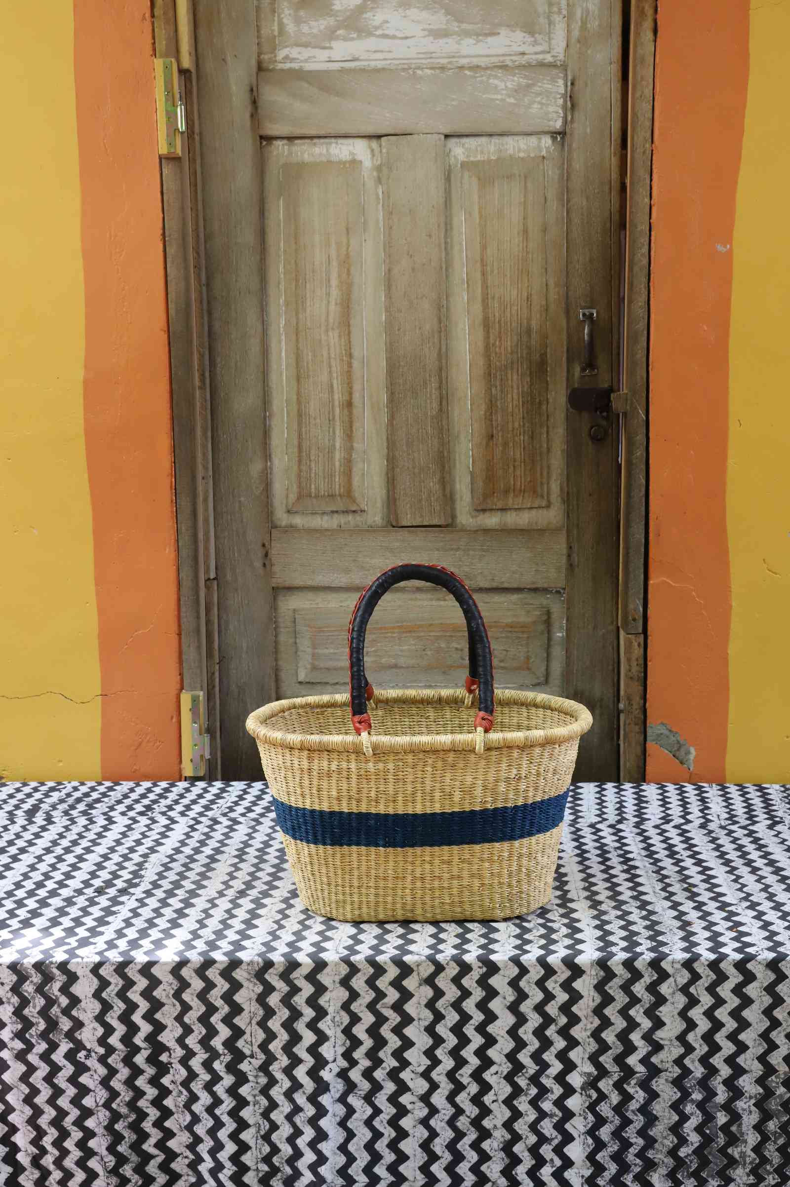 Small handwoven Bolga Zaane Tia market basket in natural straw with a navy blue band and twin leather handles.