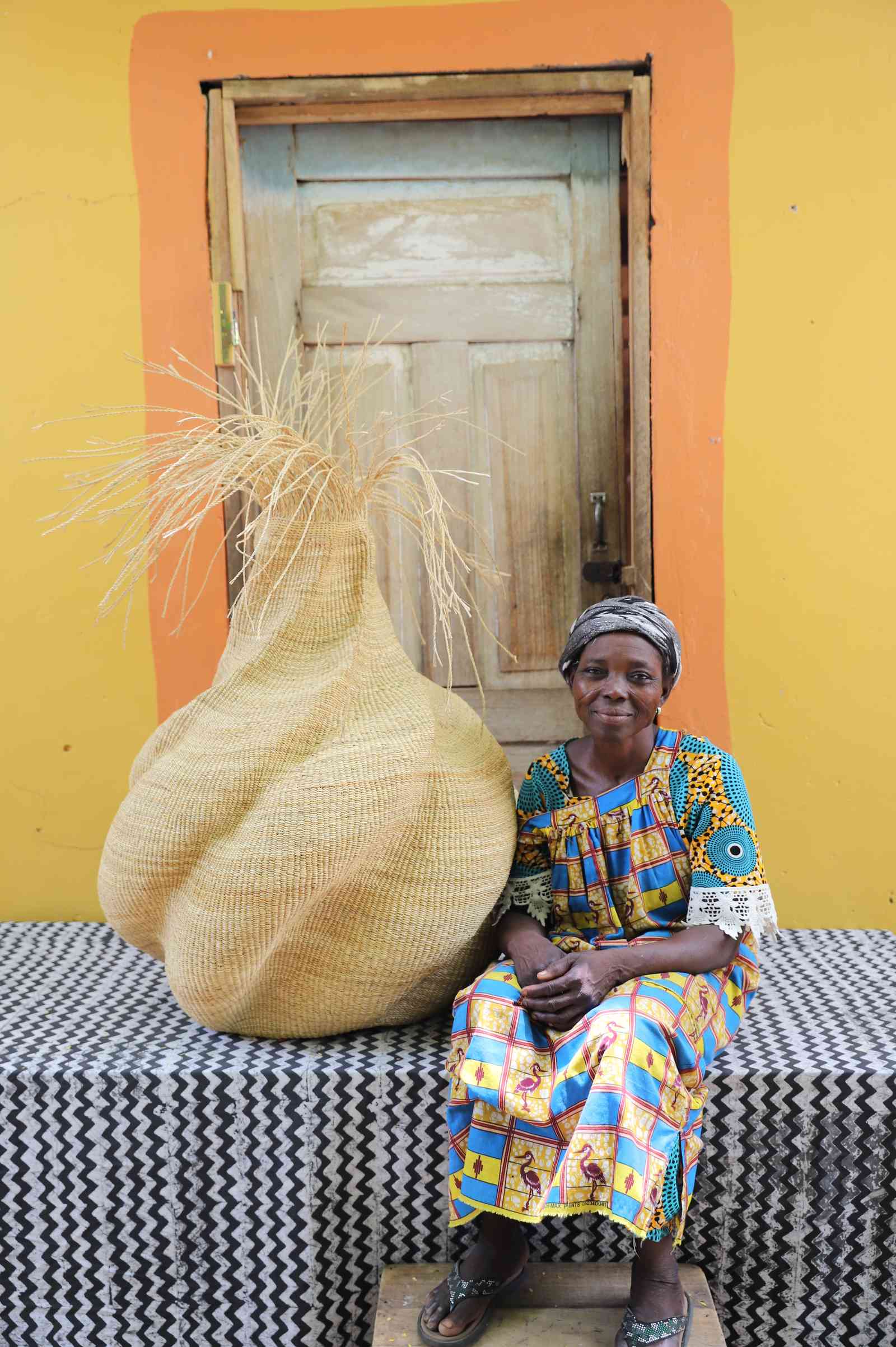 Handwoven art basket in natural straw with sculptural twisted body and dried grass stems, beside a woman in Bolgatanga.
