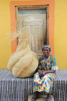 Handwoven art basket in natural straw with sculptural twisted body and dried grass stems, beside a woman in Bolgatanga.