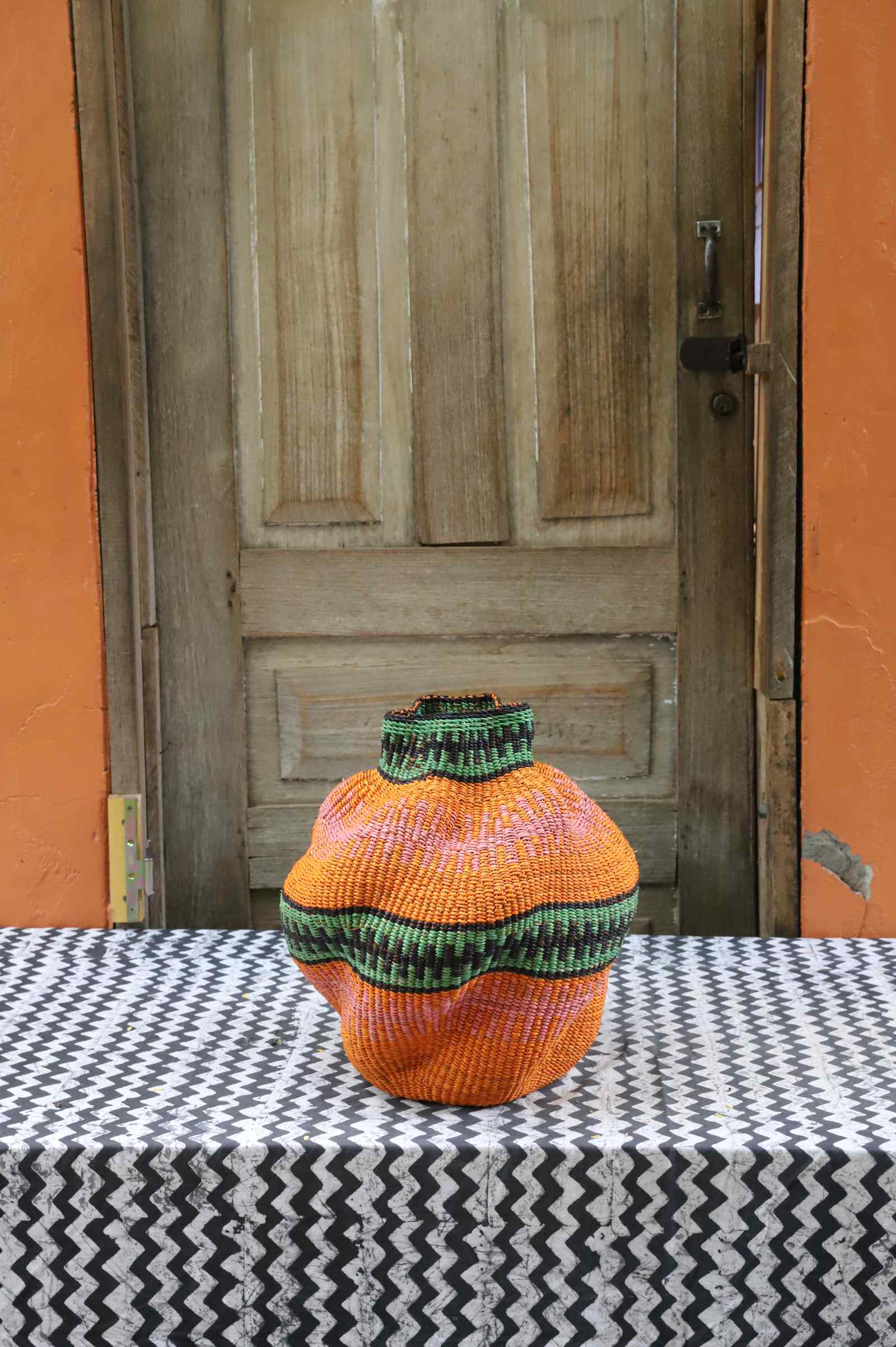 Handwoven Bolga art basket in a rounded pot shape, dyed orange with green rim and dark central band.