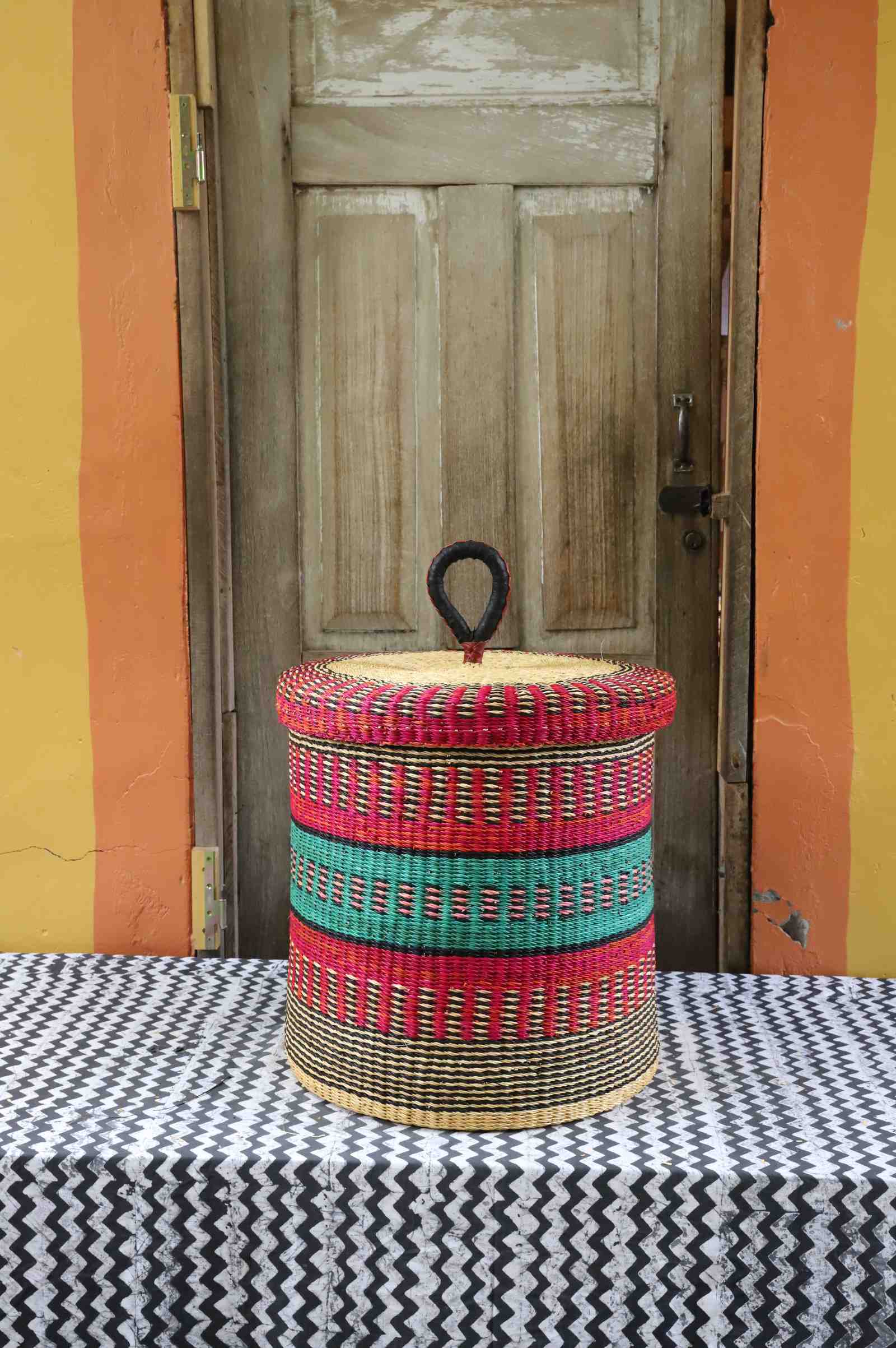 Handwoven Bolga laundry basket with lid in natural straw with bold red and green horizontal bands.