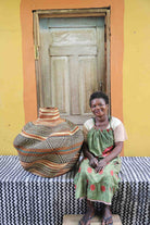 Handwoven African art basket in sculptural lobed vase form with earth-tone stripes, beside a smiling woman in Bolgatanga.