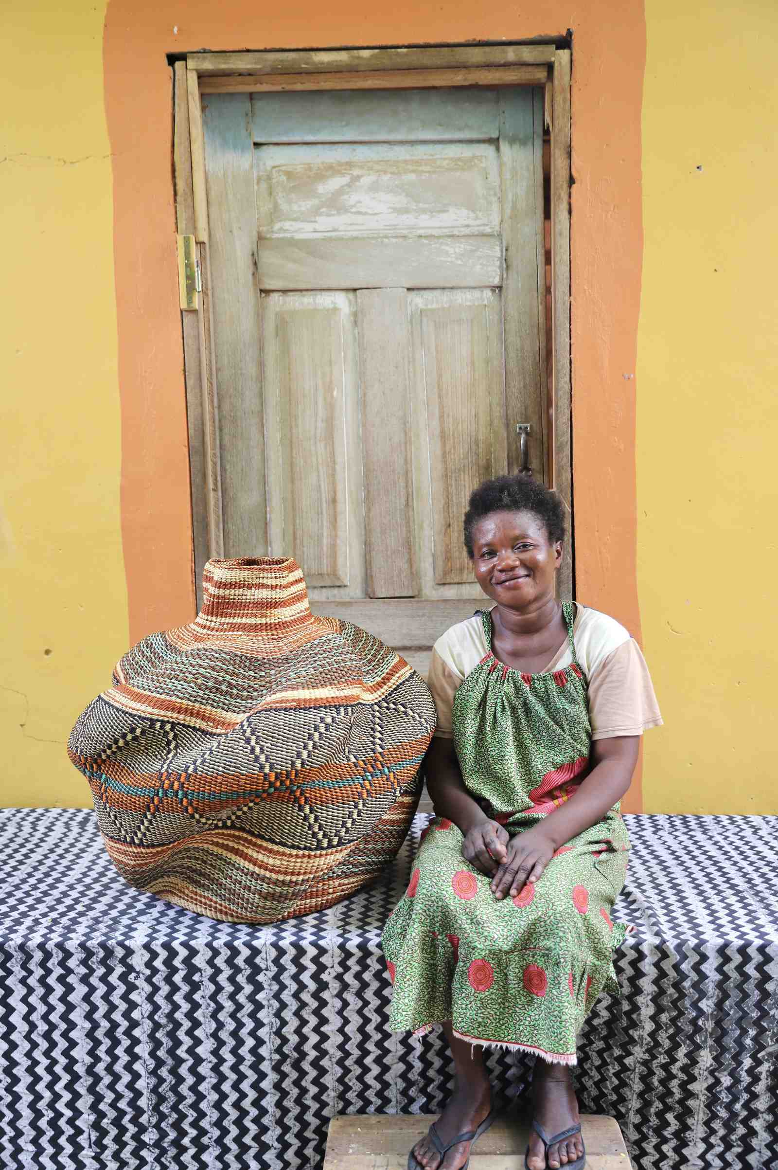 Handwoven African art basket in sculptural lobed vase form with earth-tone stripes, beside a smiling woman in Bolgatanga.