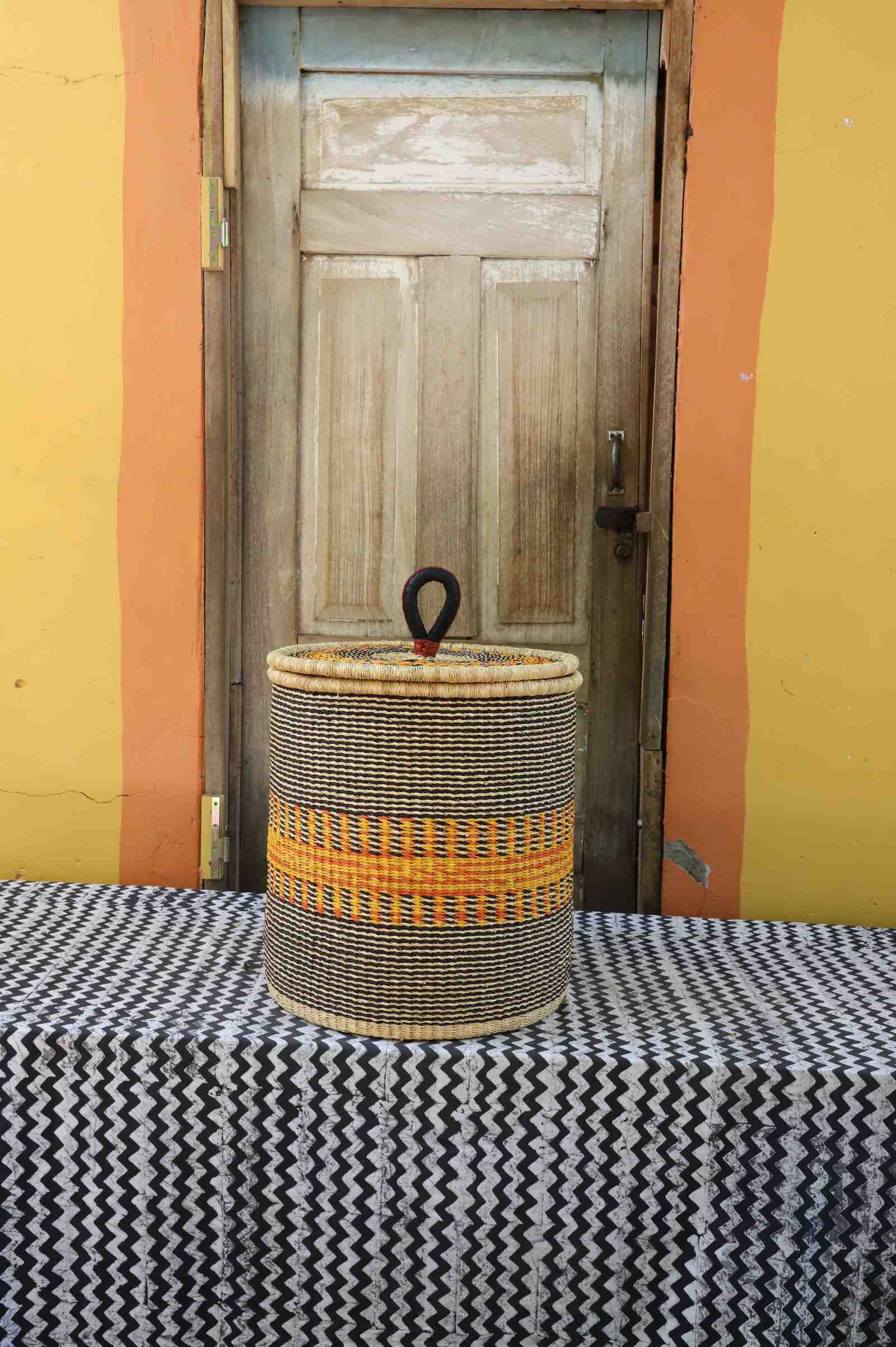 Handwoven Bolga laundry basket with coiled rim and lid in natural straw with a bold orange central band.